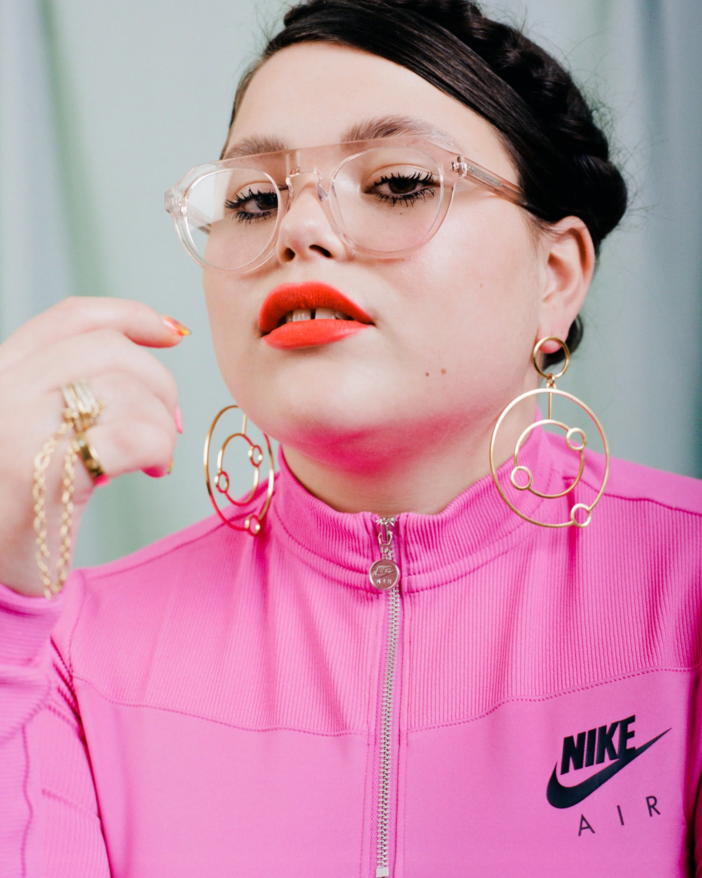 A woman with fair skin wearing clear glasses, bold red lipstick, large gold circular earrings, and a pink Nike Air zip-up jacket, looking at the camera with a confident expression.