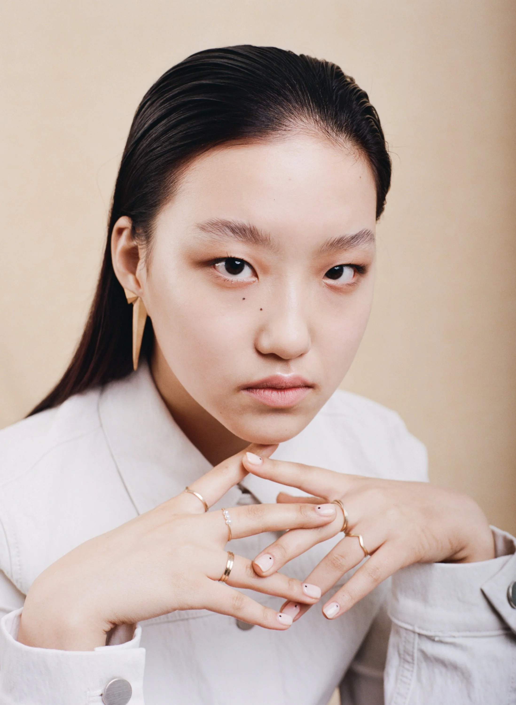 A young woman with dark, slicked-back hair, wearing earrings and multiple rings, looking directly at the camera with a neutral expression.