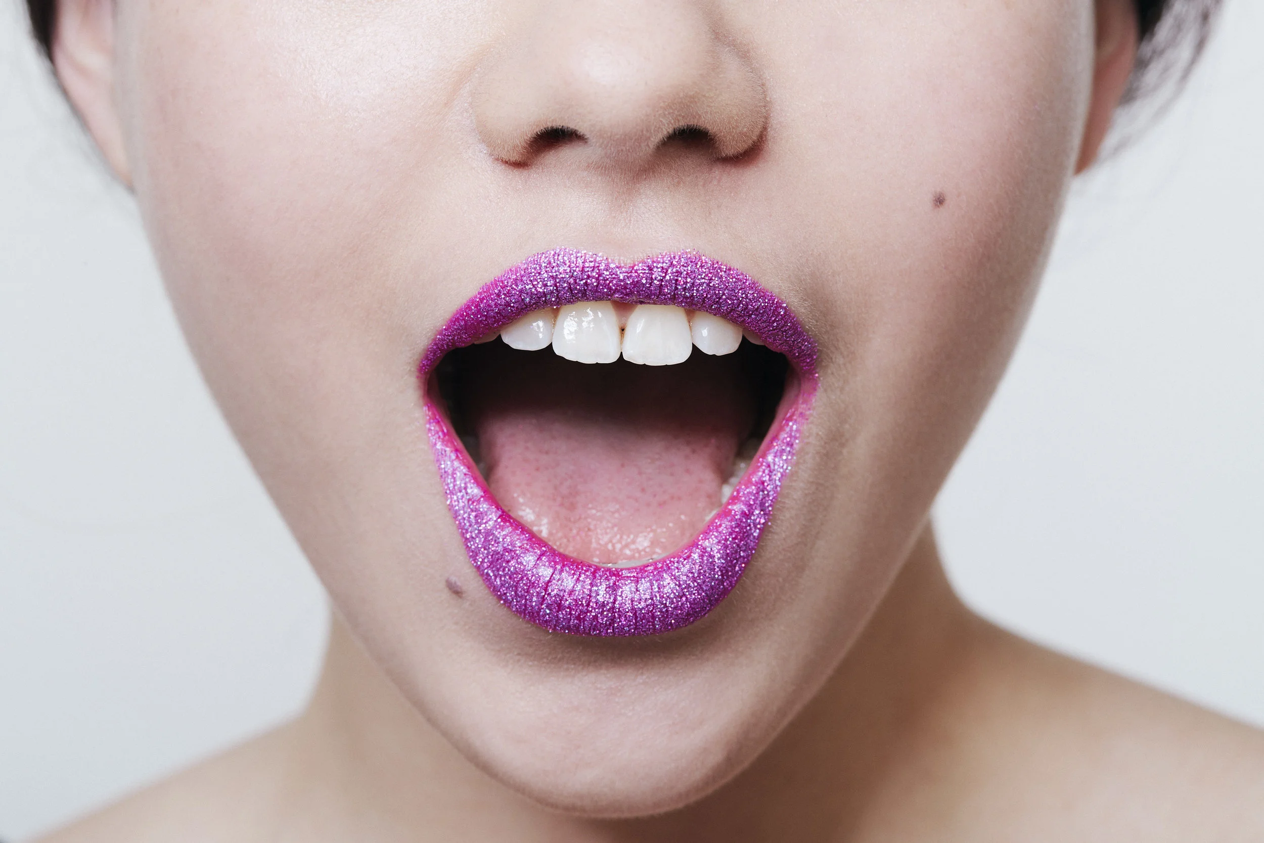 Close-up of a woman's face showing her open mouth, with lips covered in purple glitter lipstick, and teeth visible.