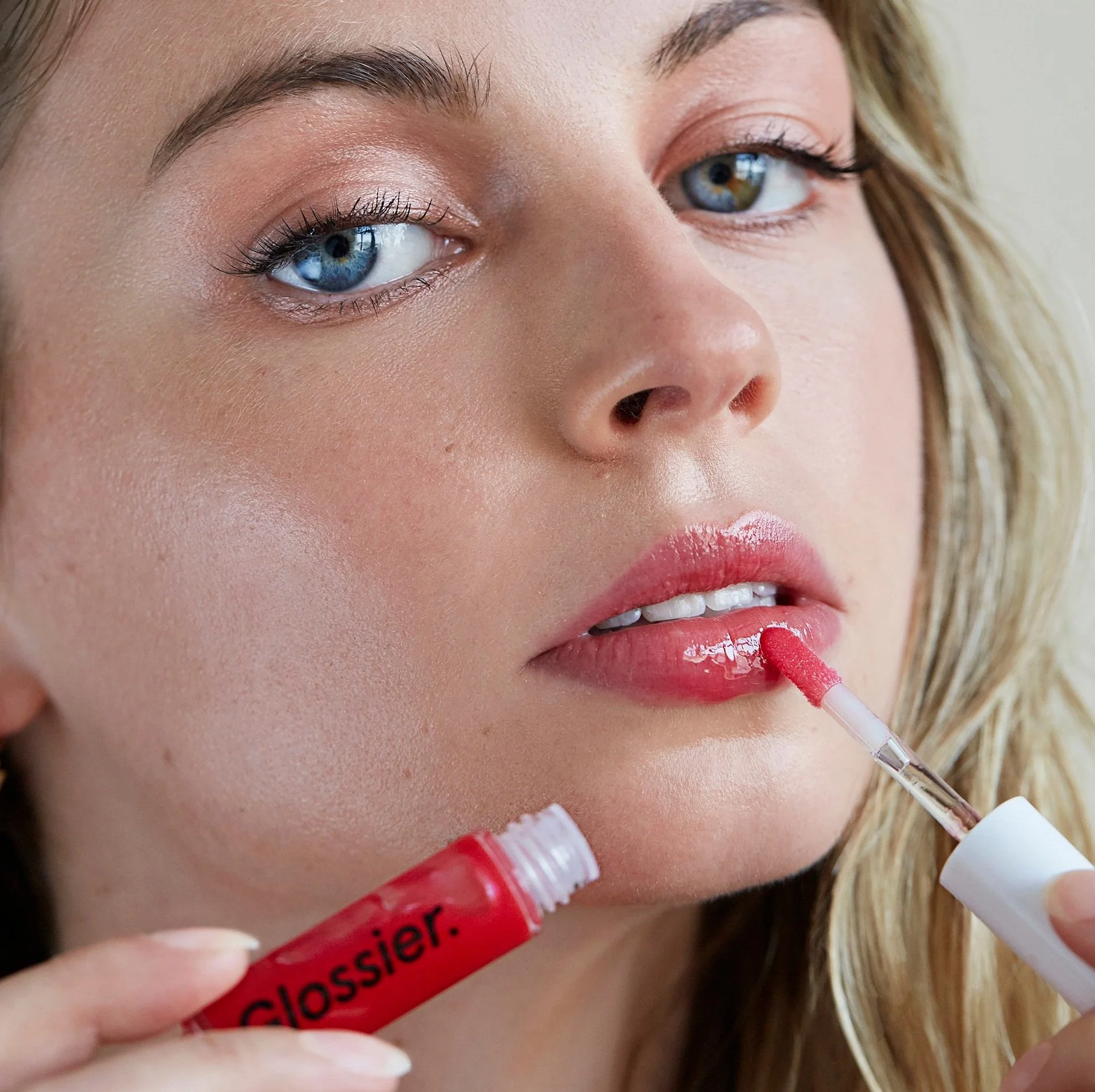 A woman with blue eyes and blonde hair applying pink lip gloss with a wand from a Glossier lip gloss tube.
