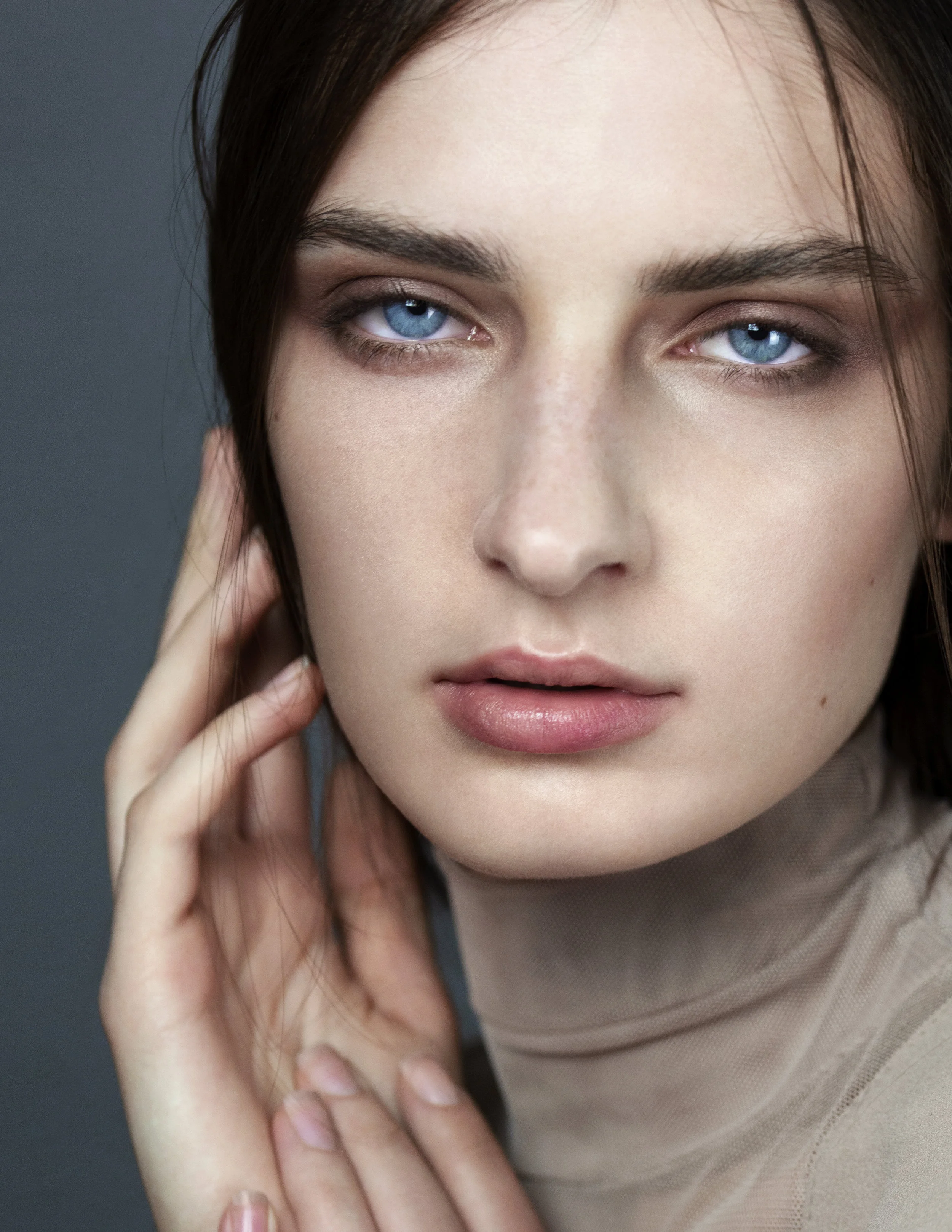 Close-up of a woman with blue eyes, fair skin, and dark brown hair, touching her face gently.