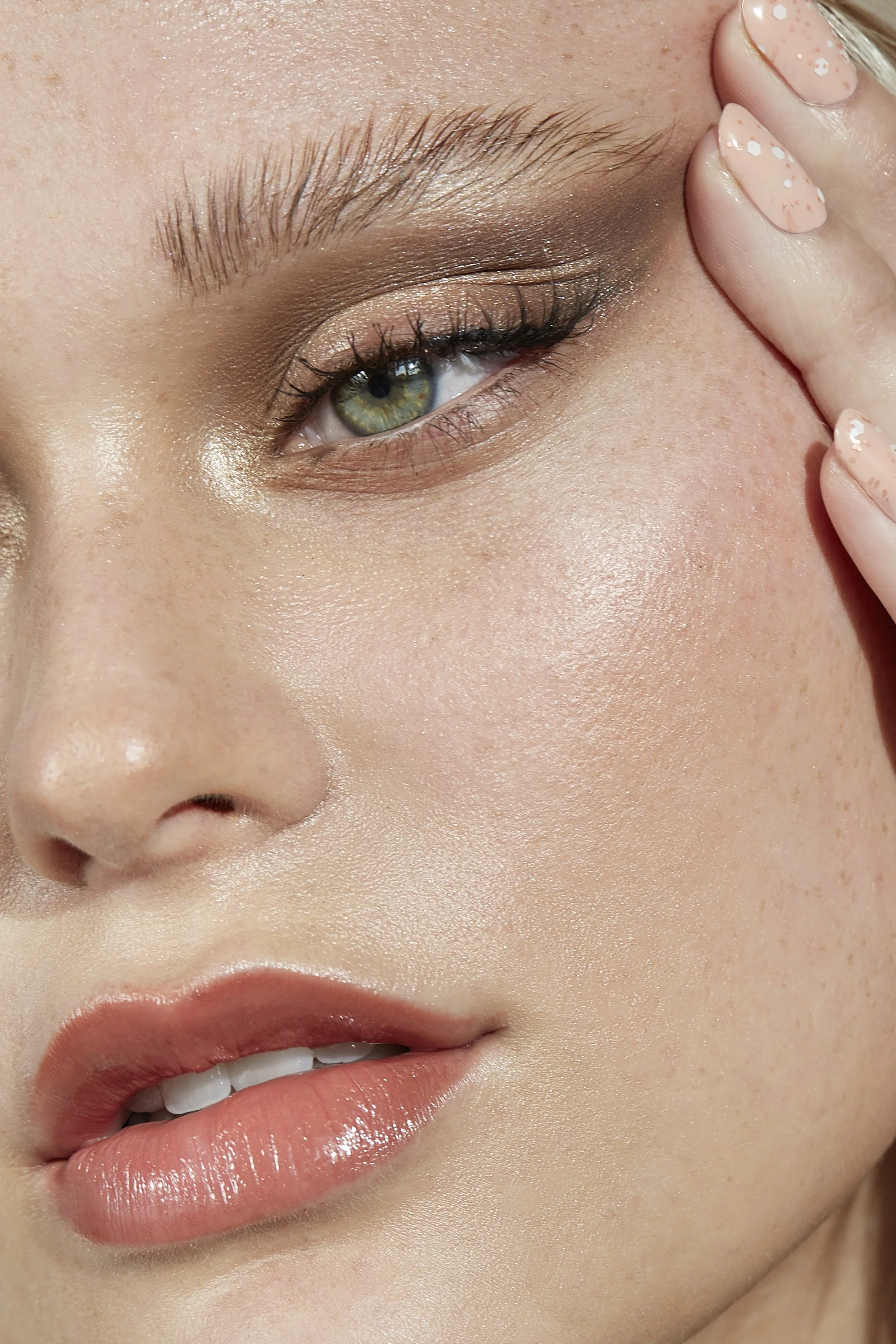 Close-up of a woman with green eyes, makeup, light skin, and peach-colored lips. She is gently touching the side of her face with her fingers.