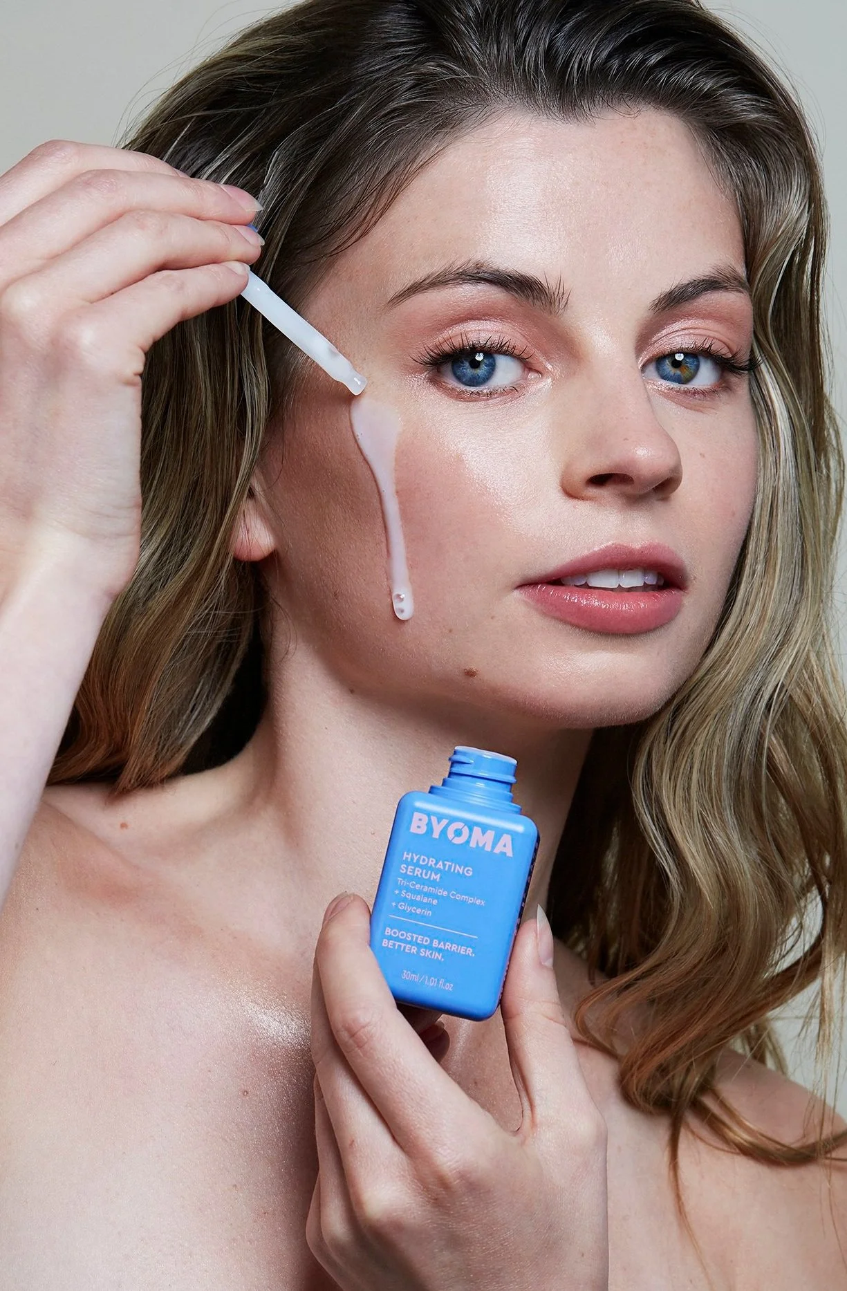 A woman with light brown hair and blue eyes applying a hydrating serum from a blue bottle labeled BYOMA to her face with a dropper.