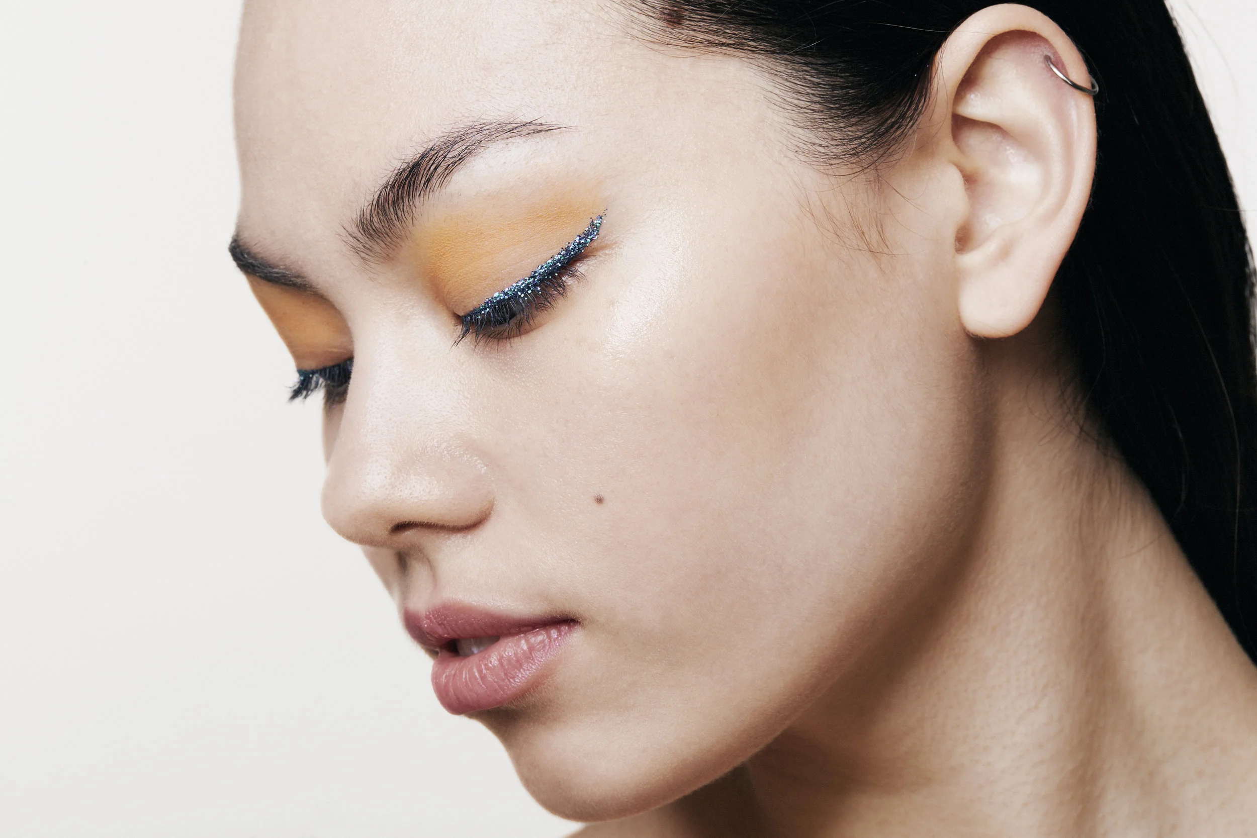 Close-up of a woman with makeup, closed eyes with yellow eyeshadow, and glittery blue eyeliner on the upper eyelids, beige lips, and a small mole above her upper lip.