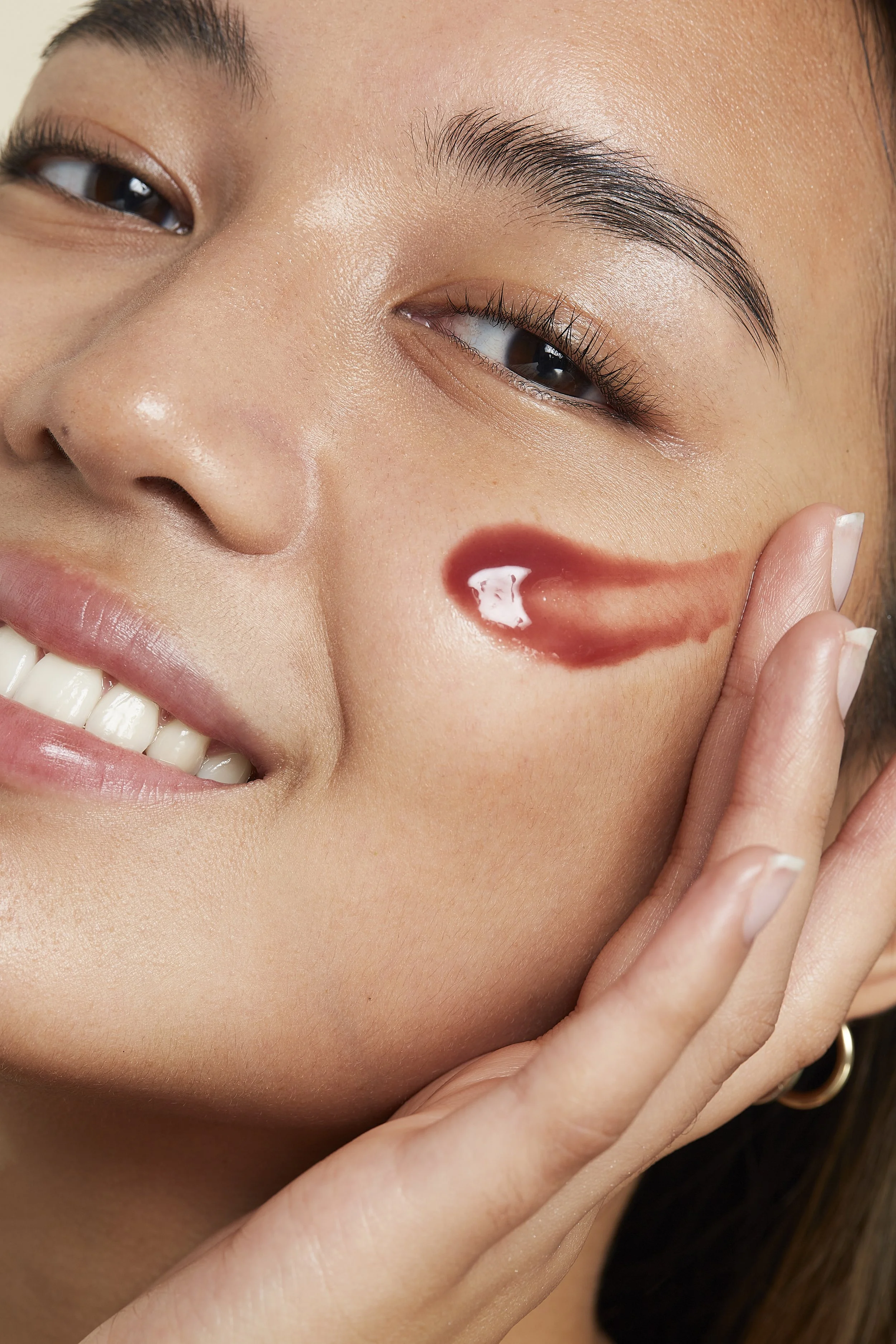 Close-up of a smiling woman with a glossy red lip stain on her cheek and a few white spots of gloss.