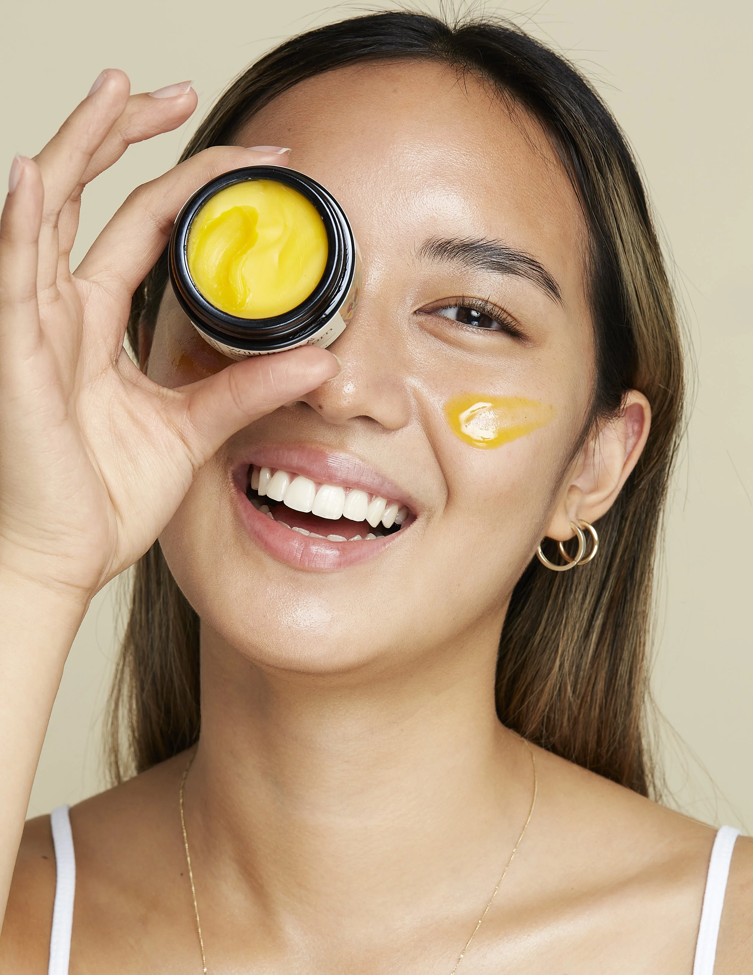 A woman with long brown hair smiling and holding a jar of yellow skincare product up to her face, with some of the product applied to her cheek.