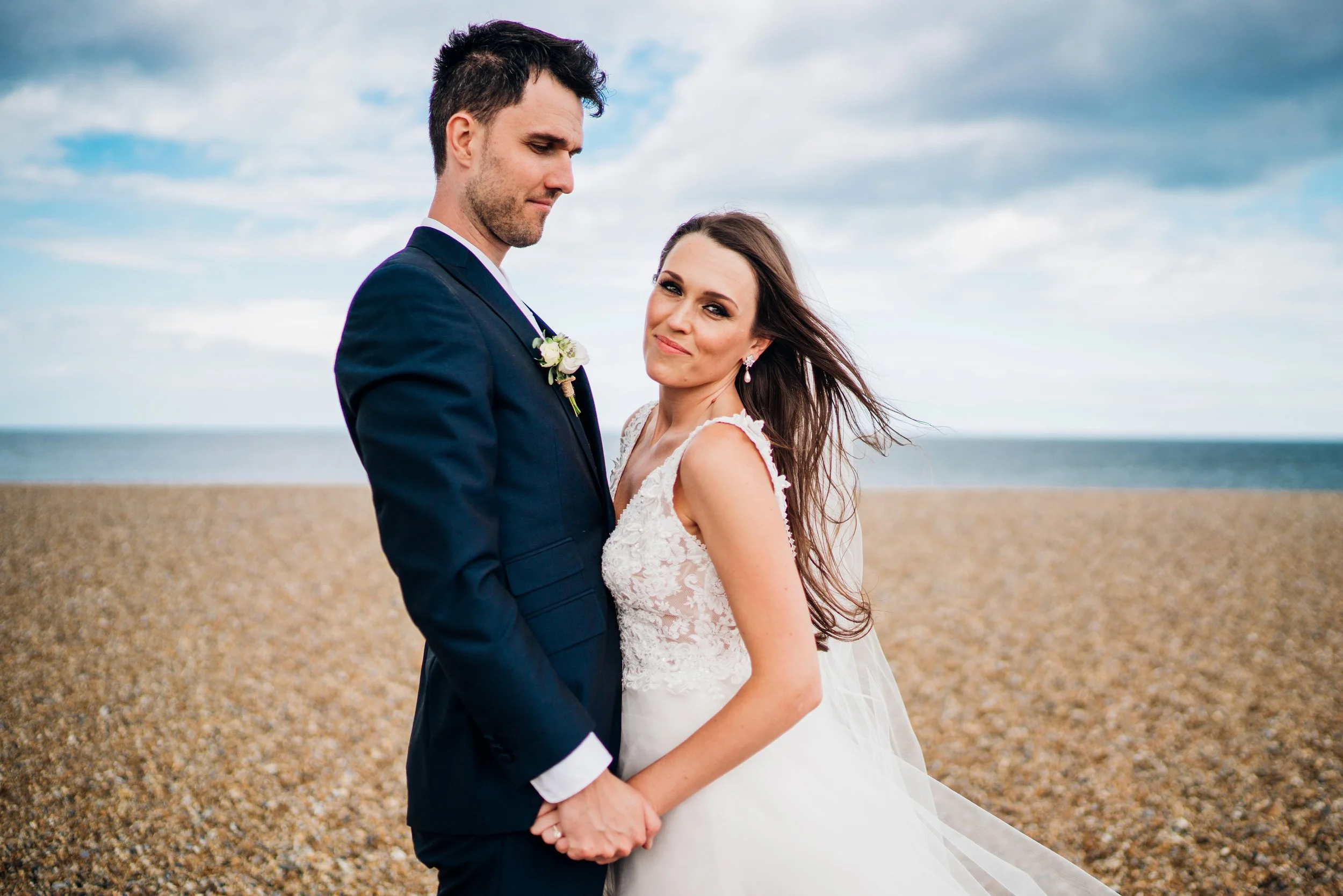 A bride and groom holding hands on the beach, with the ocean and cloudy sky in the background.