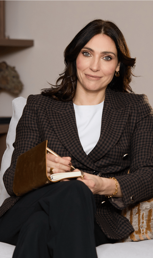 A woman sitting on a sofa holding a journal and pen, smiling at the camera, wearing a brown checked blazer, white top, and gold jewelry.