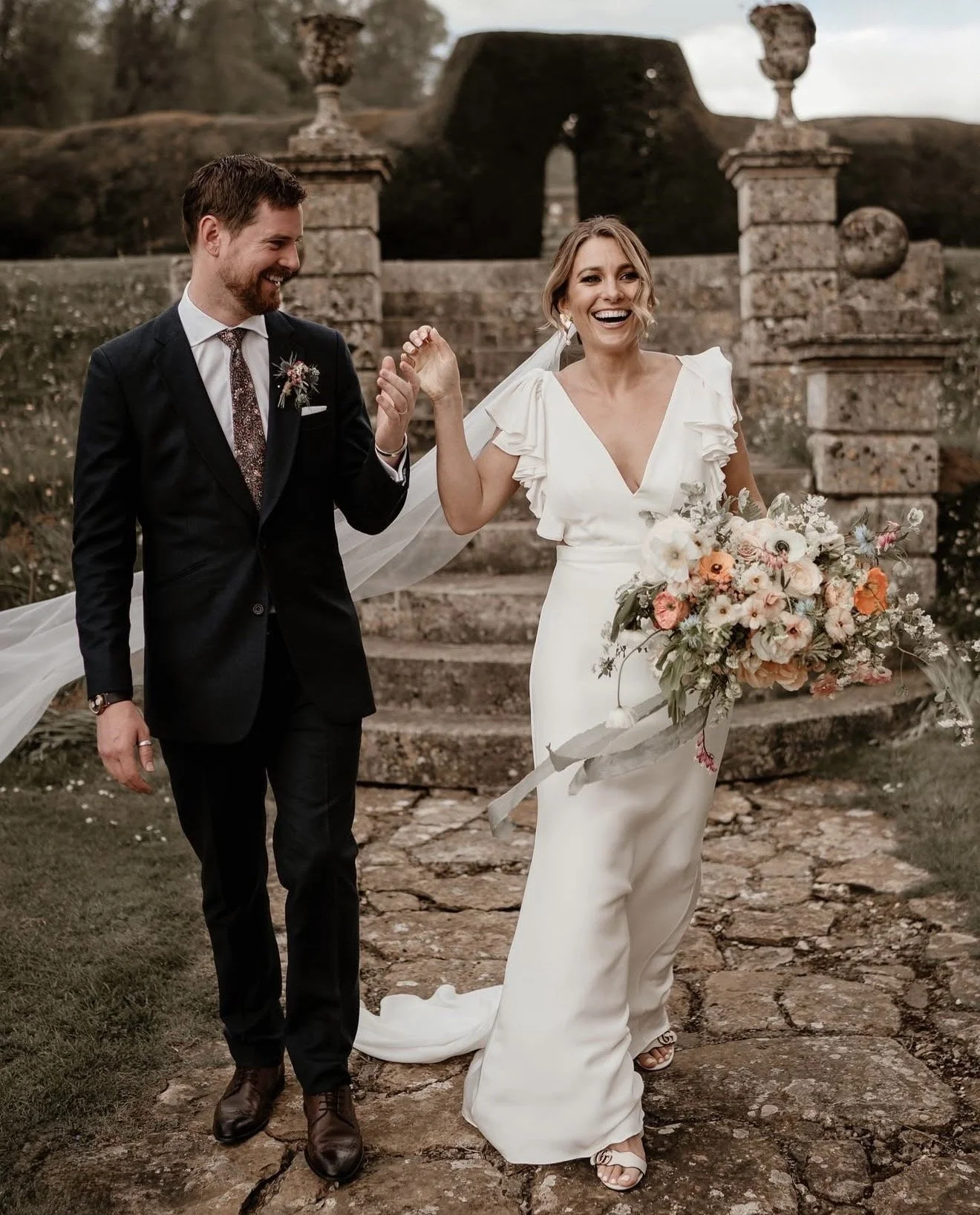 A bride and groom walking hand in hand outdoors, smiling after a wedding ceremony. The bride wears a white gown, holding a large bouquet of flowers. The groom is in a dark suit with a patterned tie. Stone steps and historic stone columns are in the background.