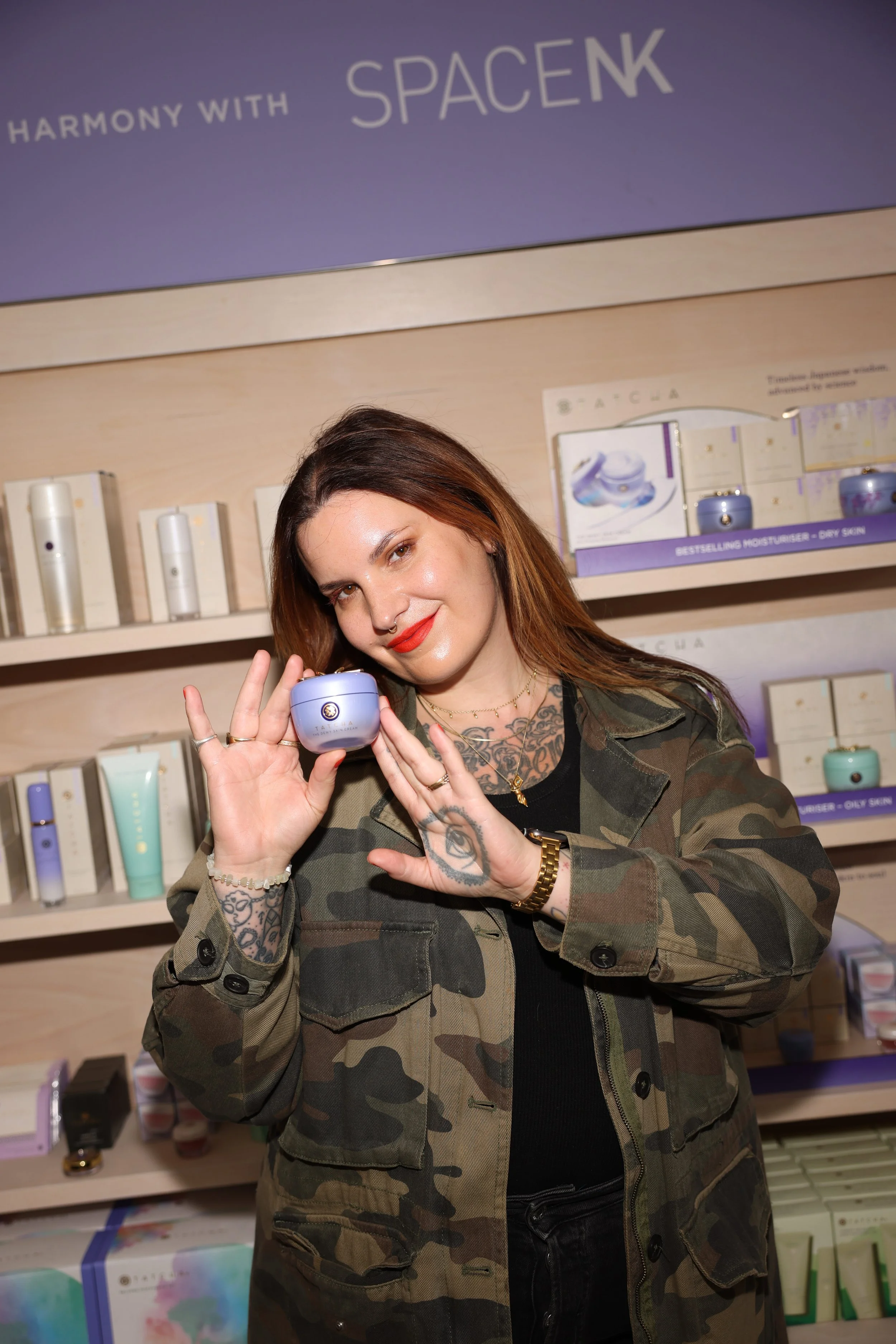 Woman with tattoos holding a purple skincare jar in a beauty store, smiling at the camera.