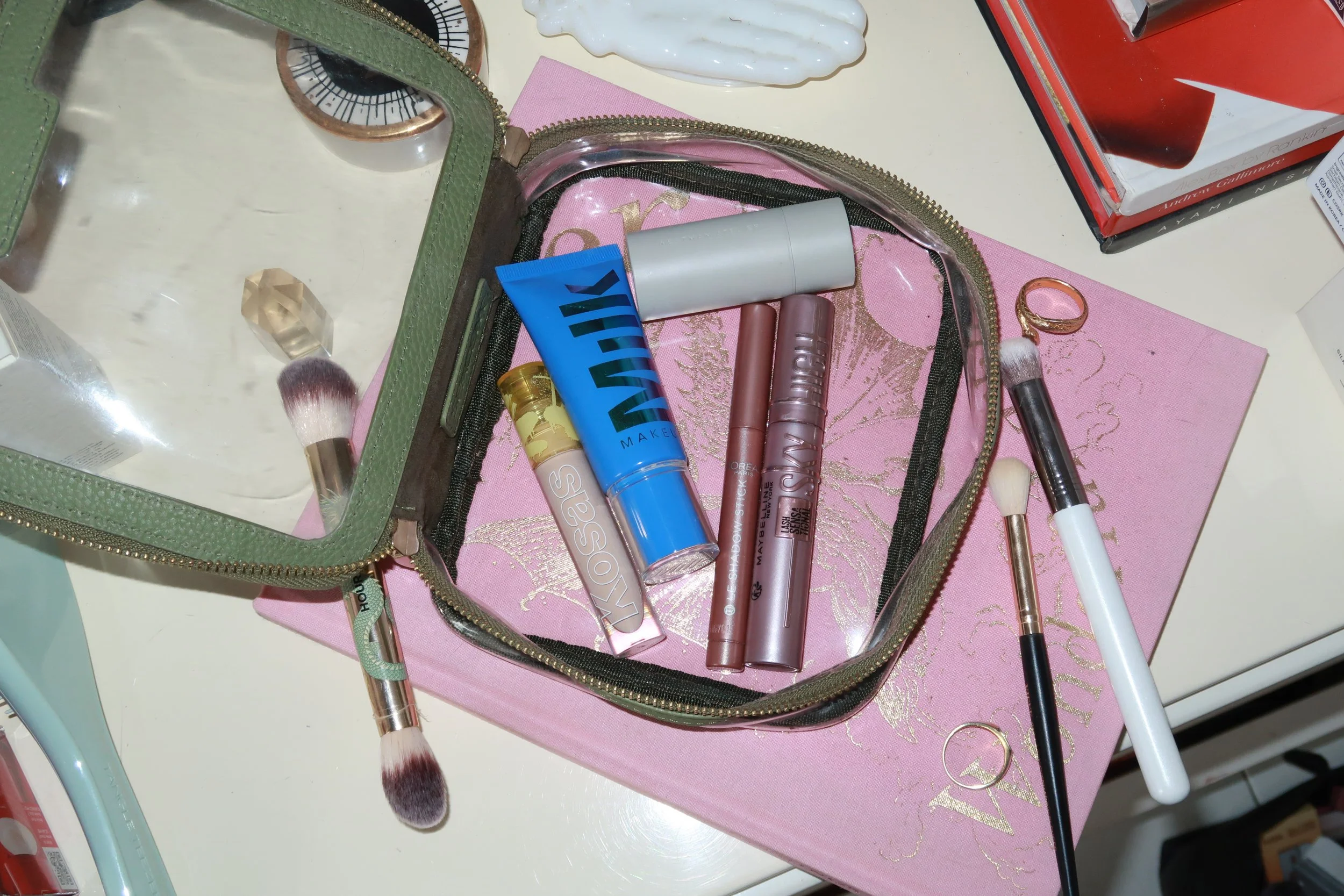 Open makeup bag containing various makeup products and brushes, placed on a pink and gold makeup palette on a white surface.
