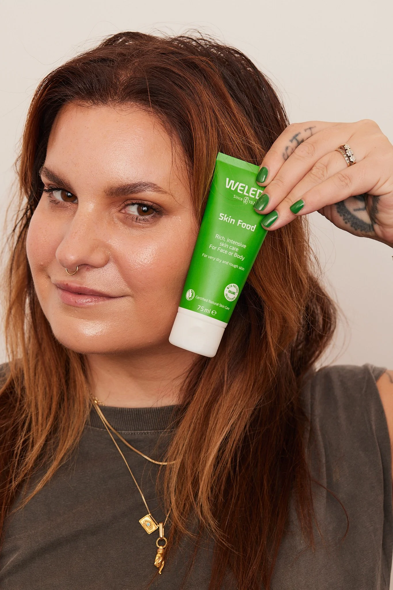 A woman with reddish-brown hair and tattoos on her hand is holding a green tube of WELEDA Skin Food near her face, smiling gently.
