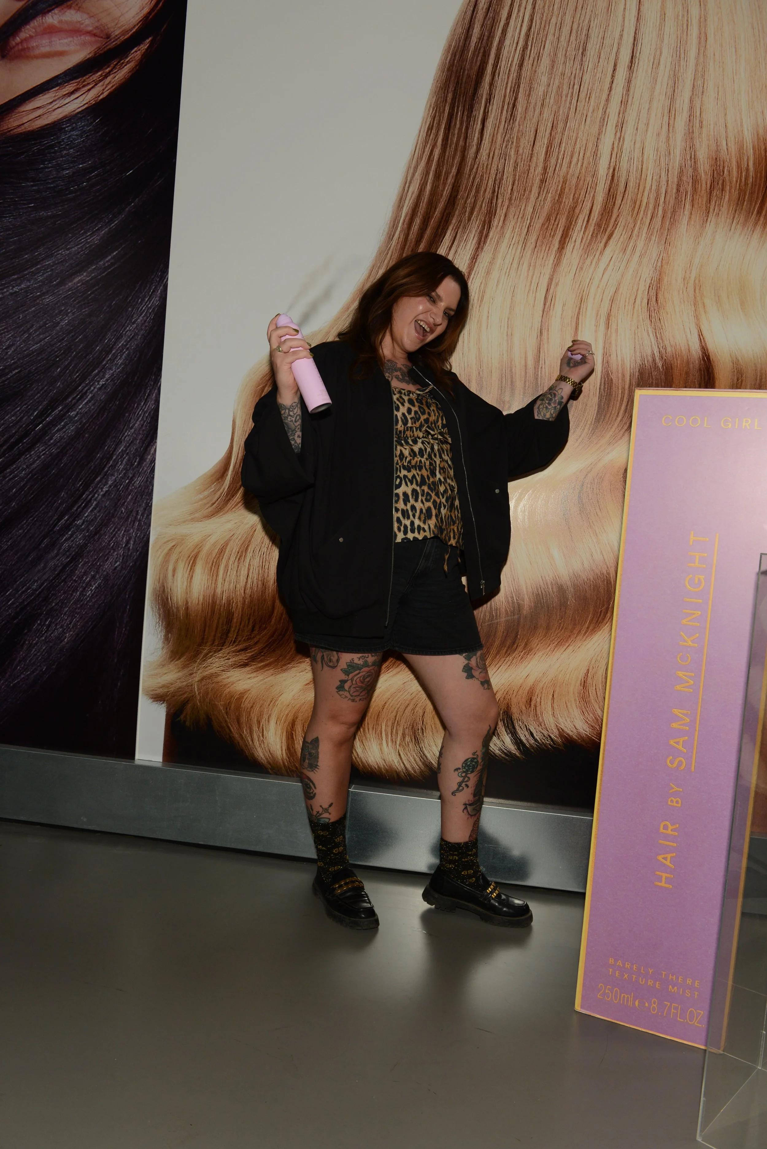 A woman with tattoos, wearing a leopard-print top, black shorts, and black shoes, standing in front of a large poster of hair, holding a pink can and posing playfully.