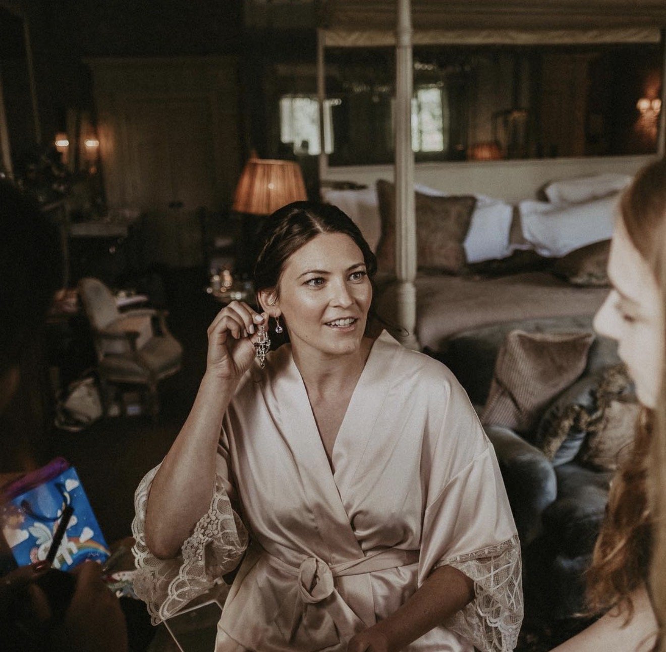 A bride in a satin robe is showing off a pair of long, dangling earrings while talking to another woman in a cozy, dimly-lit room at Babington House with a large bed and pillows in the background.