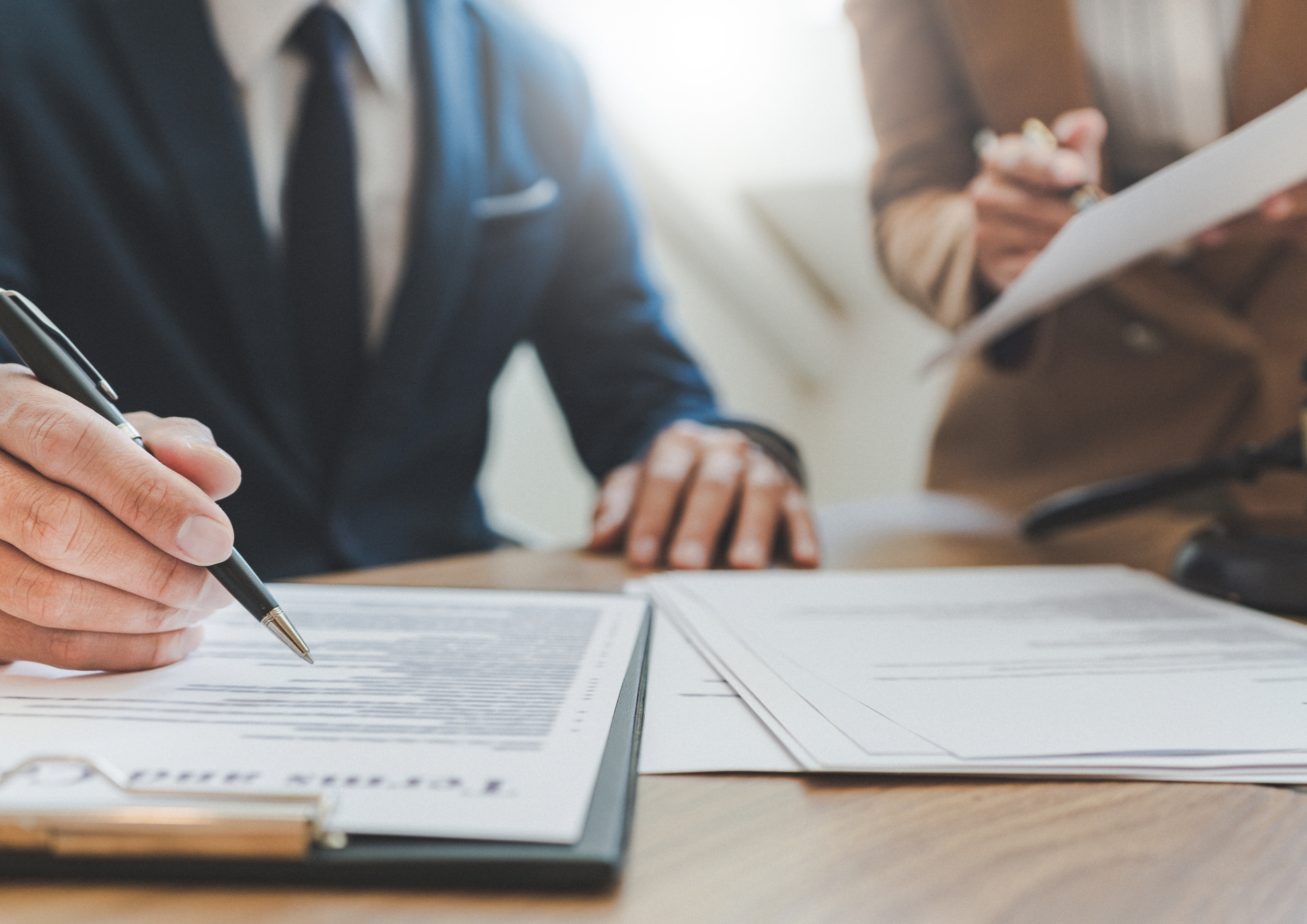 Two people in business attire reviewing documents at a desk, with one person writing with a pen on papers and the other holding a pen and a sheet of paper.