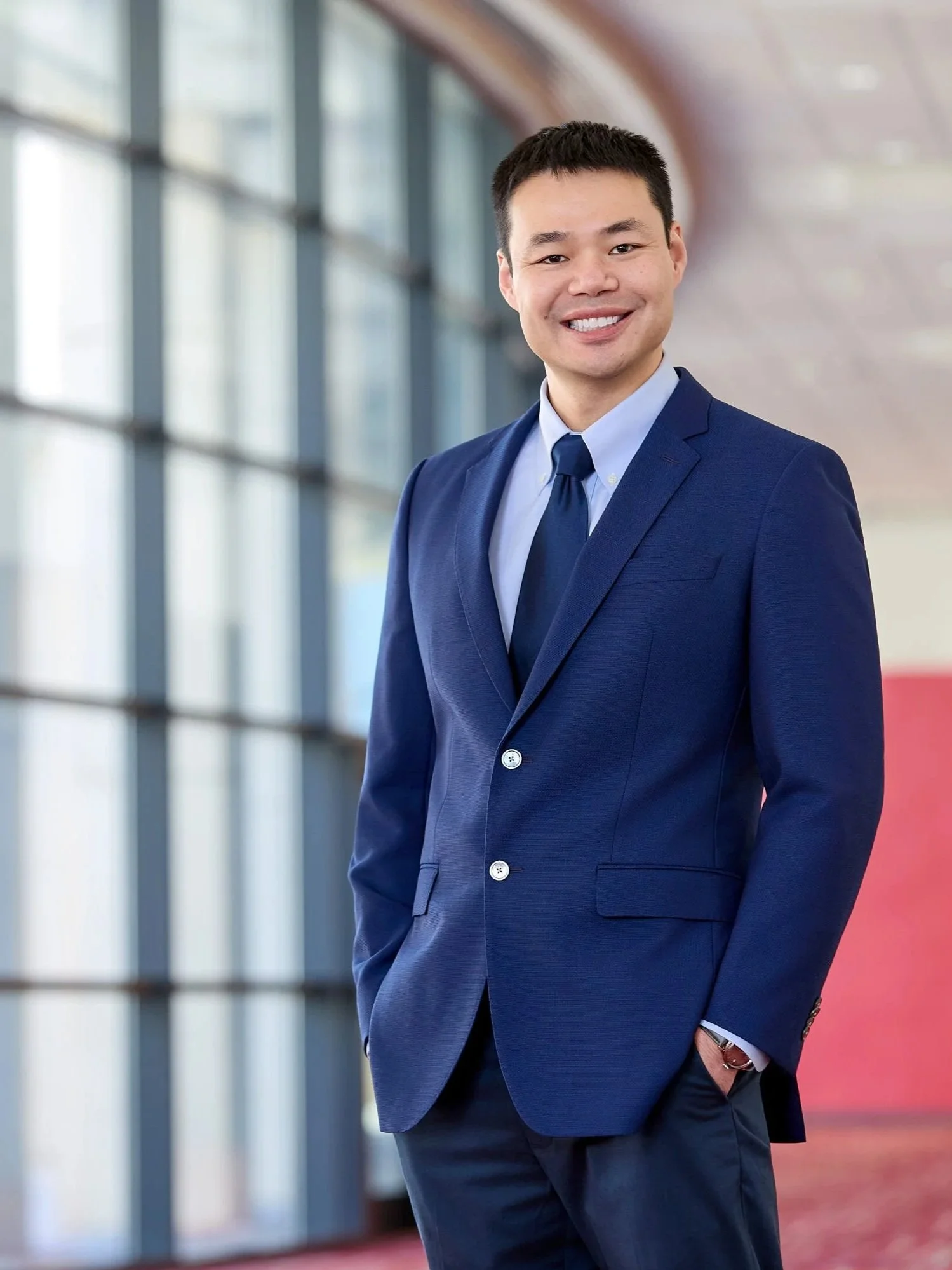 A smiling Asian man in a navy blue suit and tie stands with his right hand in his pocket in a modern building with large windows.