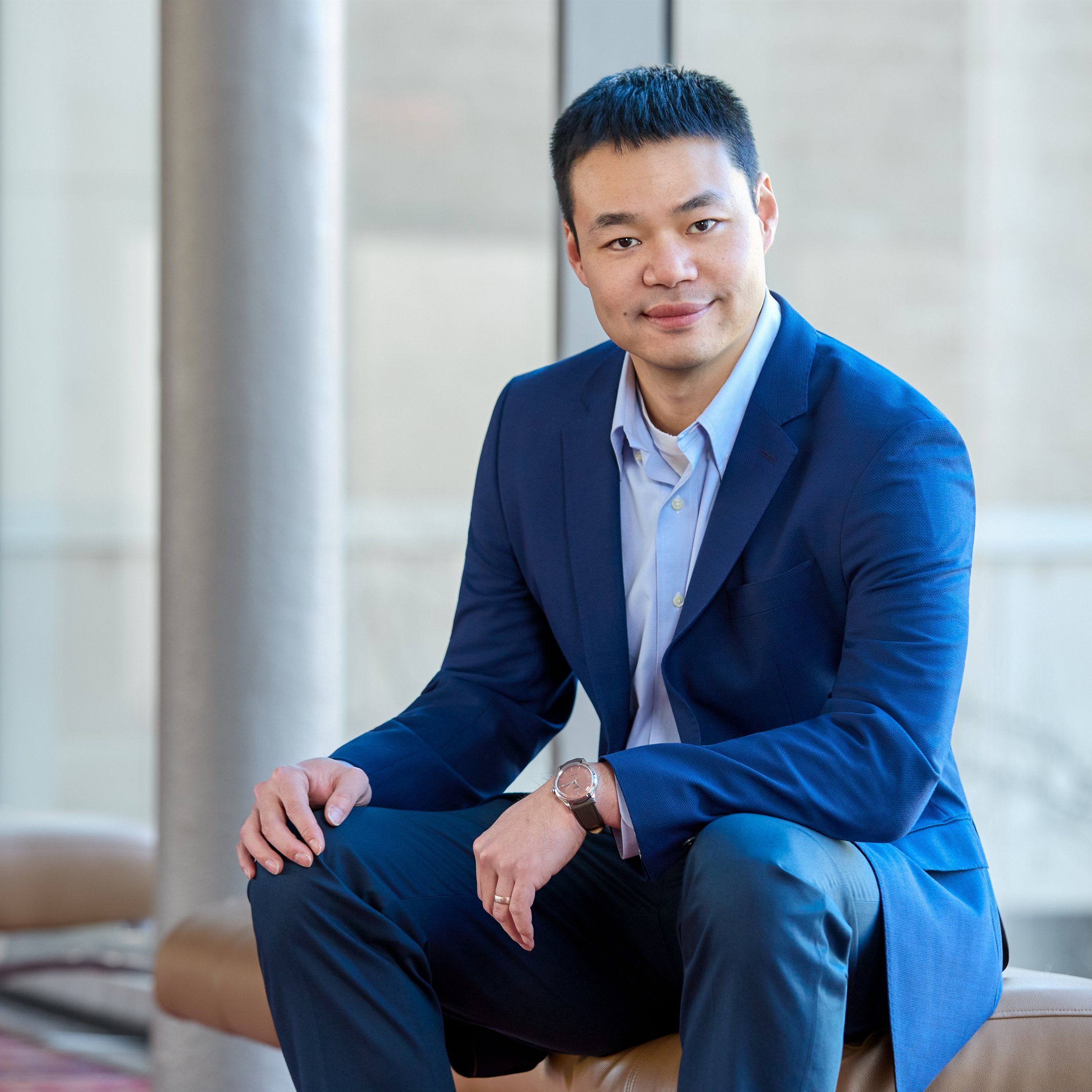 A young Asian man in a blue suit and light blue shirt sitting on a bench in an indoor setting, smiling at the camera.