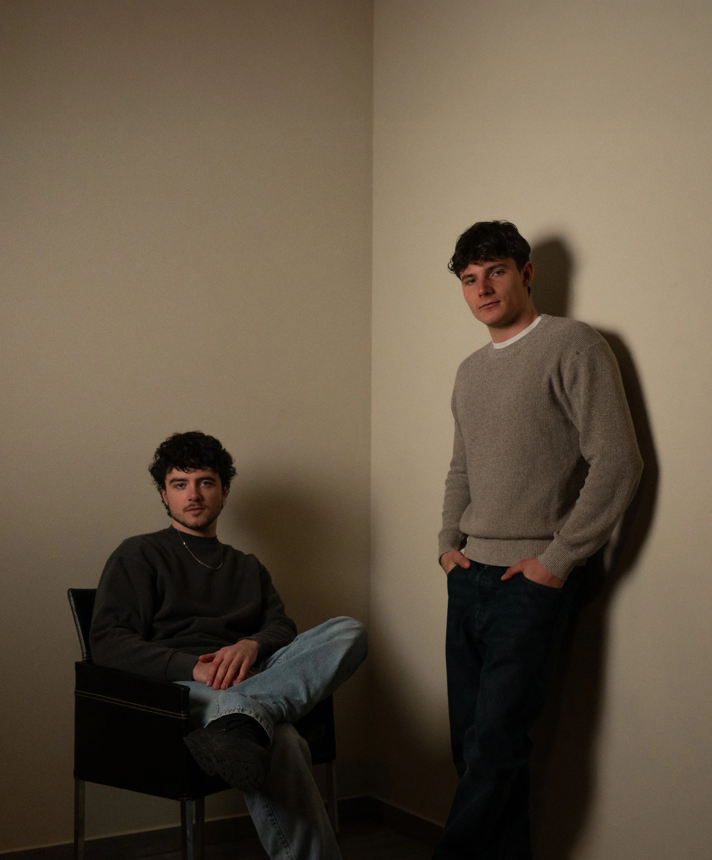 Zwei junge Männer in einem Raum mit weißen Wänden, einer sitzt auf einem Stuhl, der andere lehnt an der Wand.