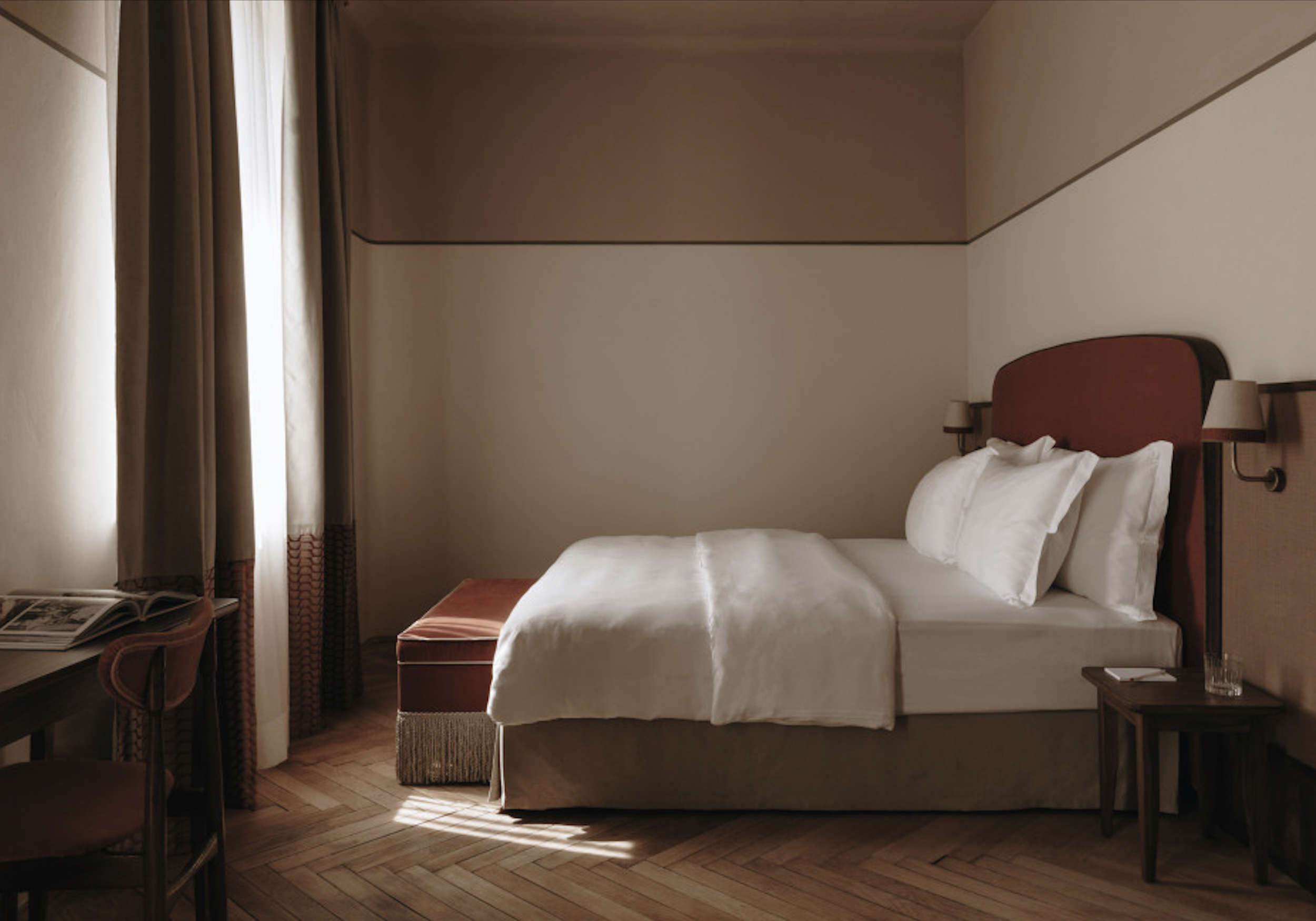 Ein einzelnes Bett in einem minimalistisch eingerichteten Hotelzimmer mit einem Kopfteil in Rot, weißen Bettwäsche und einem Fensterschirm aus dicken Vorhängen, die Licht hereinlassen.