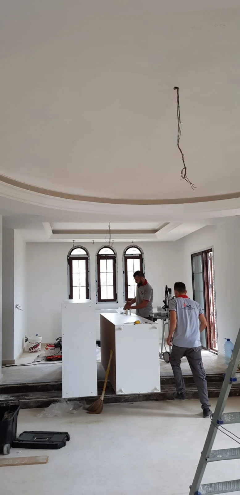 Two workers are installing or assembling kitchen cabinets in a room with large arched windows and a tray ceiling. One worker is standing behind the cabinet, and the other is walking towards it, with construction tools and materials scattered around.