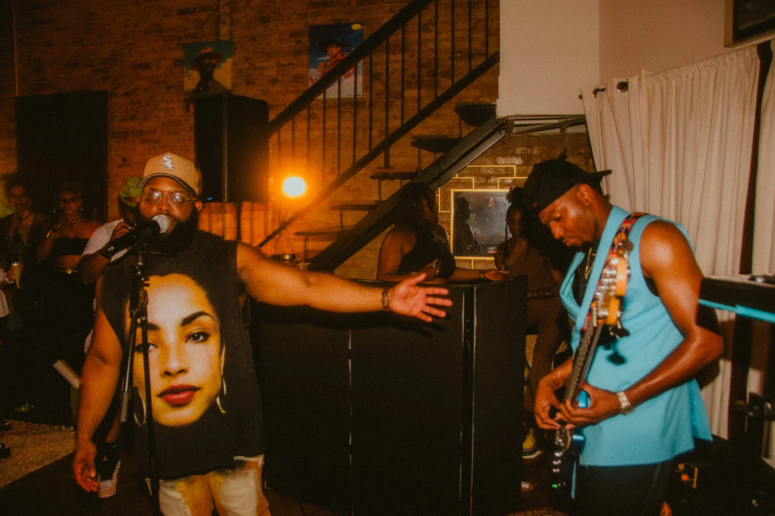 A lively indoor gathering with people socializing and dancing. The room has exposed brick walls decorated with posters of animated characters. In the foreground, a person wearing a blue dress and a black cap is playing a guitar. A large poster of a w