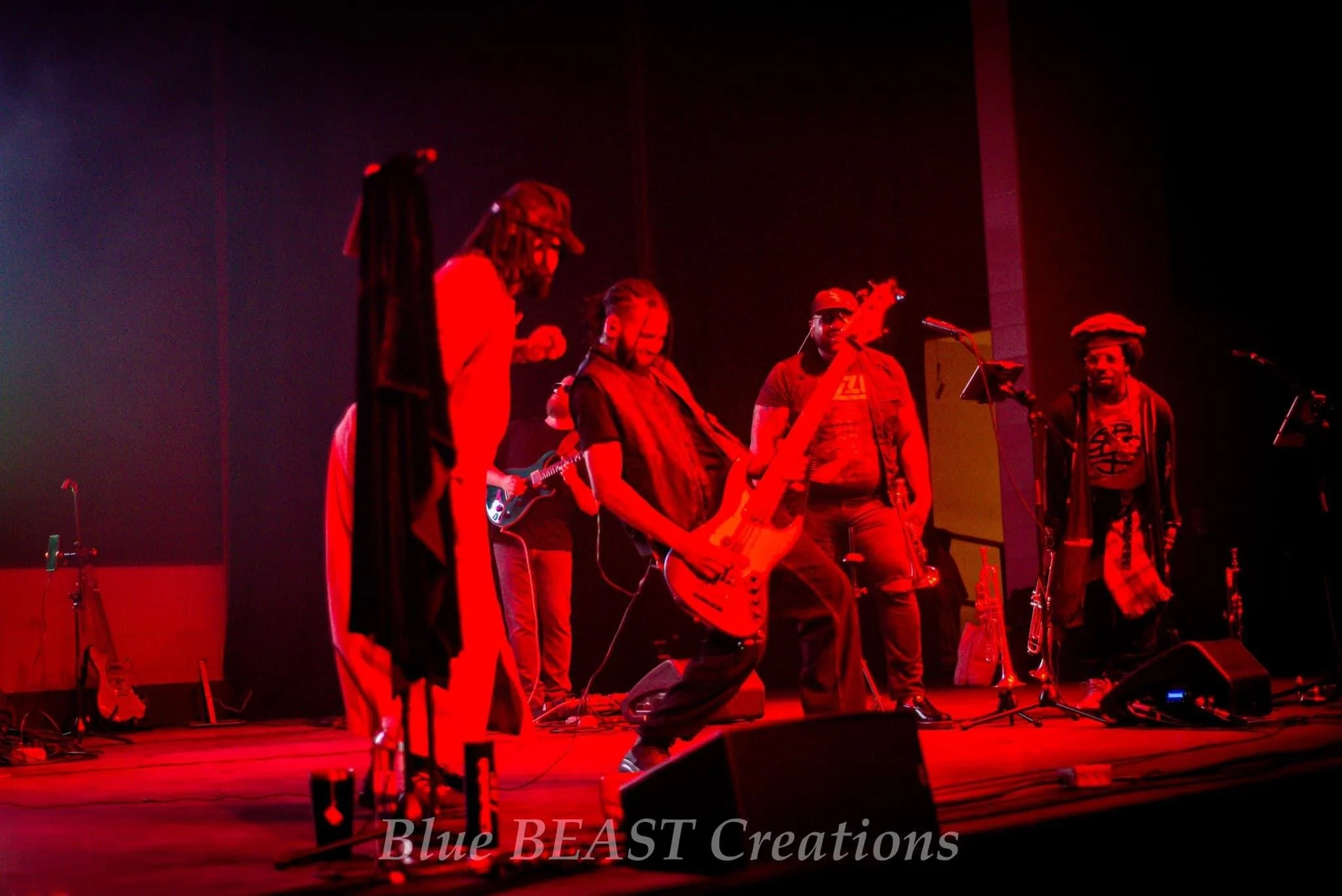 Musicians performing on stage under red lighting, including a man with dreadlocks playing an electric guitar, others singing and playing instruments, microphones, and music stands visible.