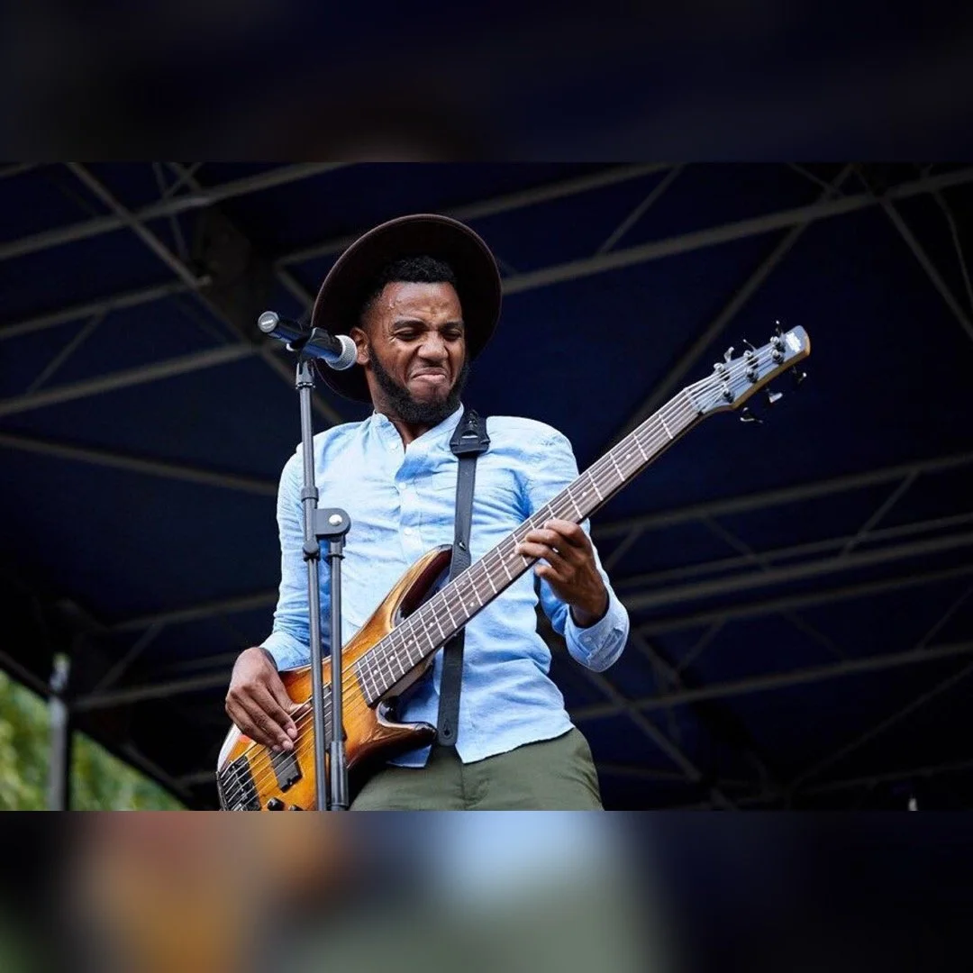 A man playing an electric bass guitar on stage with a dark background, wearing a light blue shirt, a wide-brimmed hat, and making an expressive face.