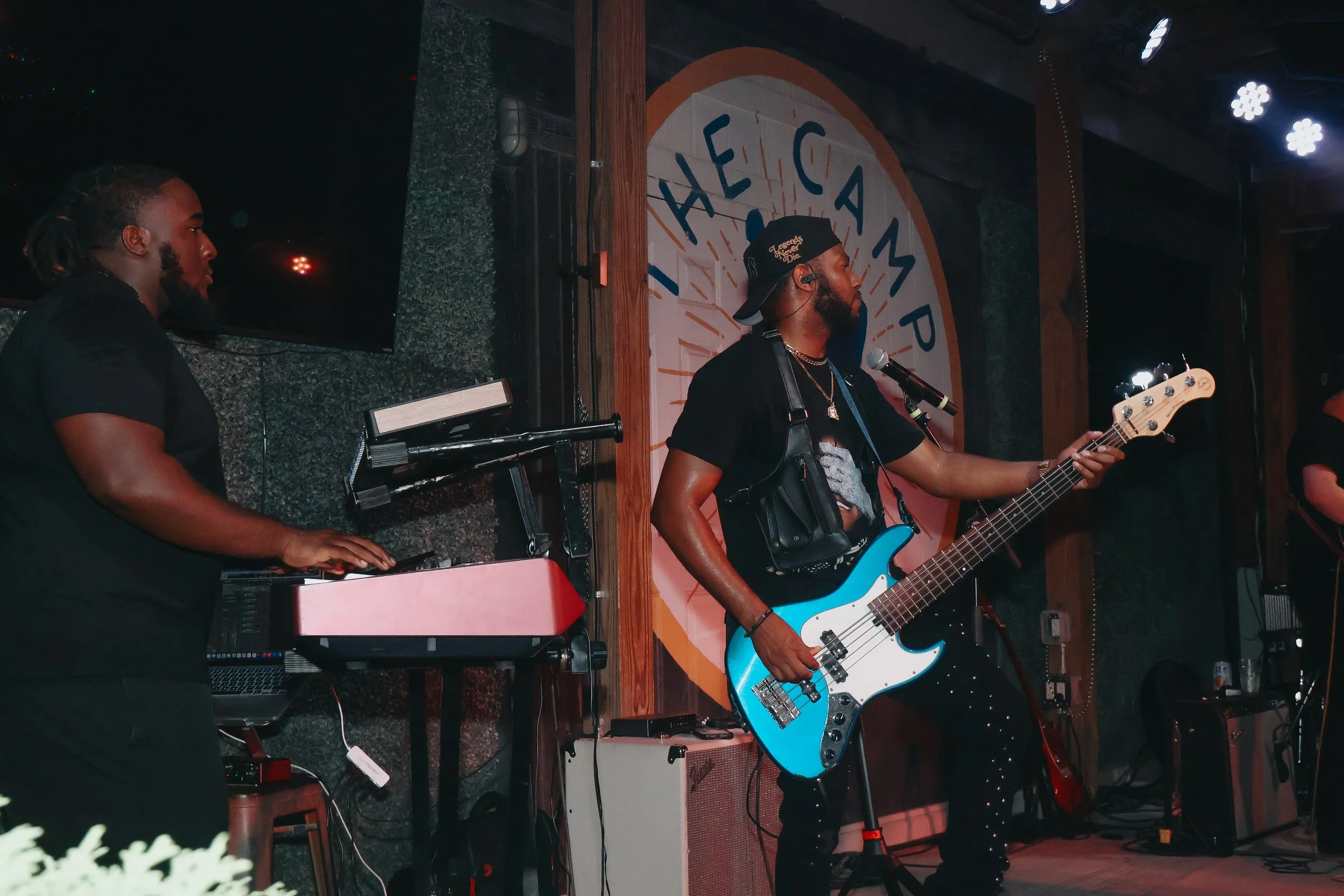 A band of two musicians performing on stage: a keyboard player and a bass guitarist, with a large sign reading 'Hale G' in the background and stage lighting overhead.