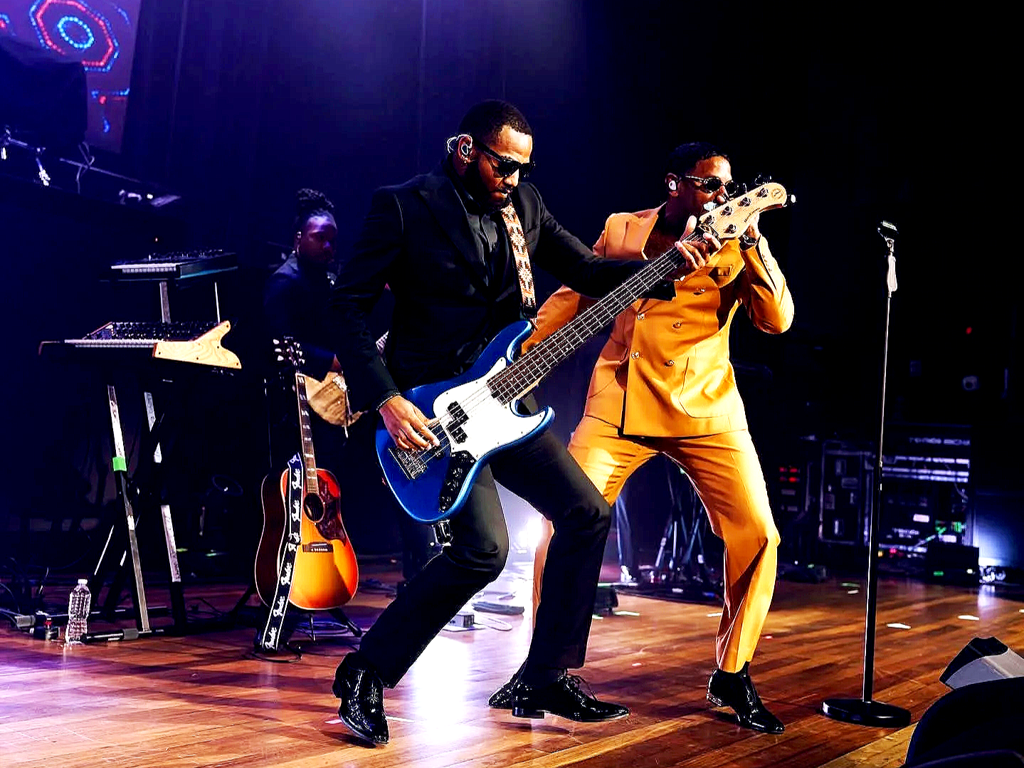 Two musicians playing electric guitars on stage during a concert. One man is wearing a black suit and sunglasses, playing a blue electric guitar. The other man is dressed in a bright yellow suit, also wearing sunglasses, and playing a bass guitar. In the background, there is a keyboard and other musical equipment.