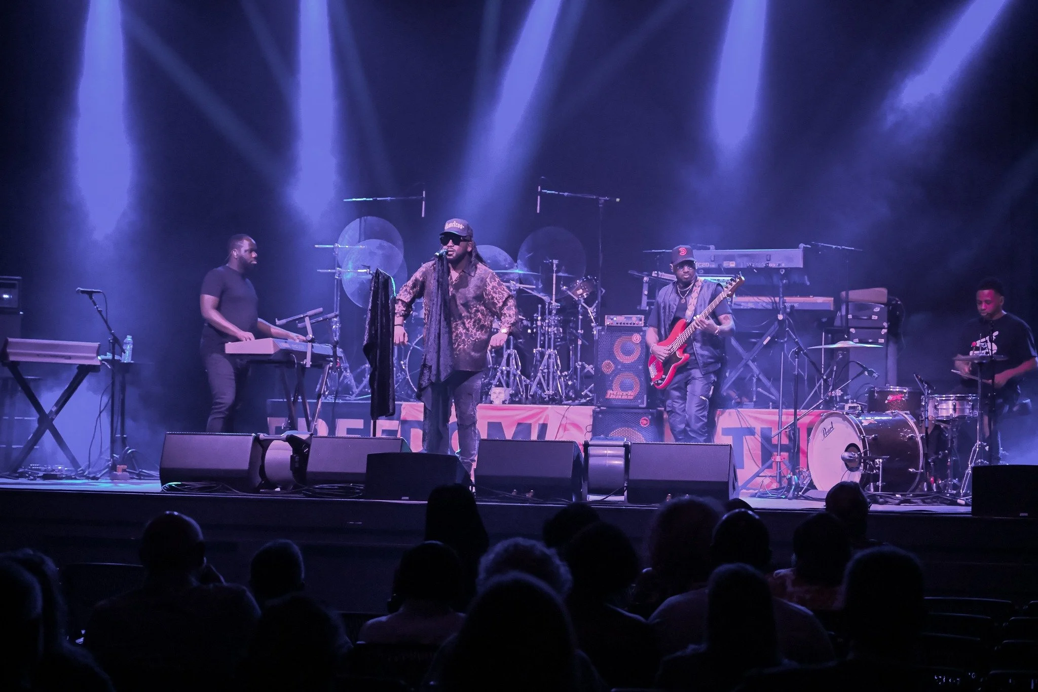 A band performs on stage with five musicians under purple and blue stage lighting, with audience members visible in the foreground.