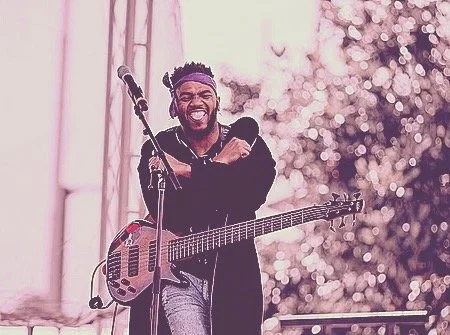 A man with dreadlocks and a beard smiling while holding an electric guitar during a performance with a microphone in front of him, outdoors with a blurred background of trees and lighting.