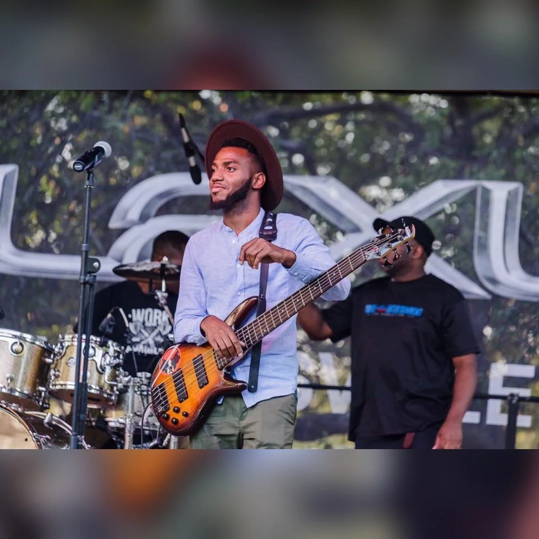 A group of musicians performing outdoors, including a man in a hat holding a bass guitar and another man playing drums in the background.