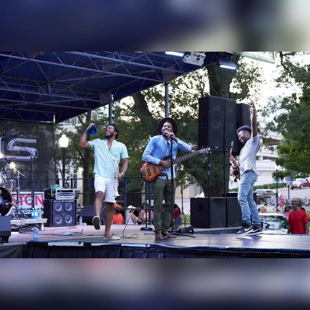 Three musicians performing on an outdoor stage, with a person singing and another playing the saxophone, in front of an audience during the evening.