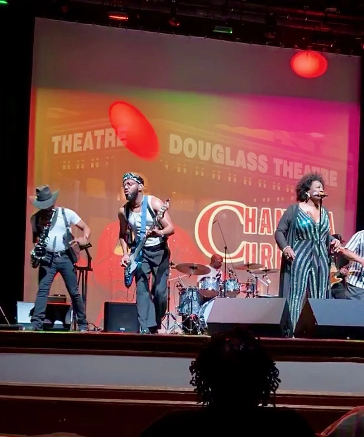 A musical performance on stage with four musicians, including a drummer, guitarist, saxophonist, and vocalist. The background displays a colorful screen with the words 'Doughlass Theatre' and 'Charly Smart.' The stage is lit with red and green lights