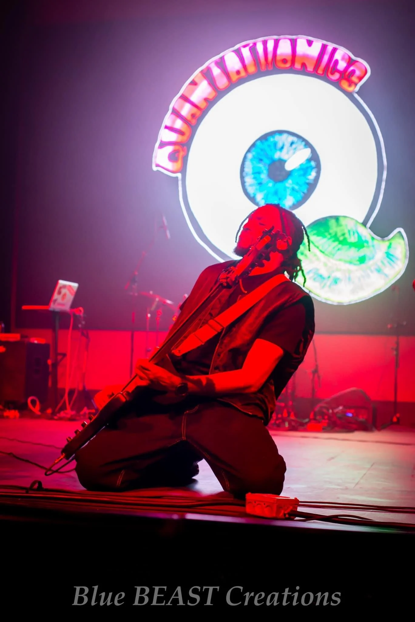 A person kneeling on stage playing a guitar, with vibrant neon signs in the background, including a large eye and a stylized rainbow-colored shape, and the text "Blue BEAST Creations" at the bottom.