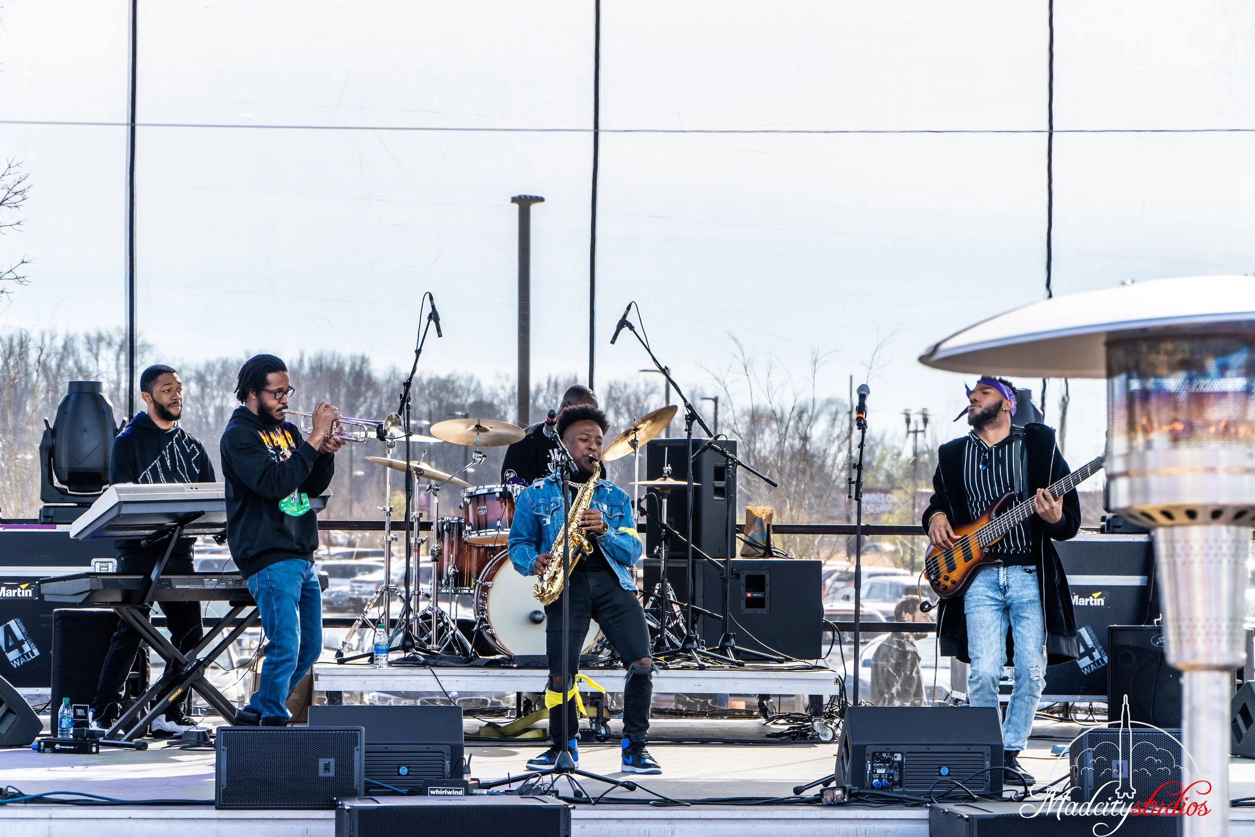 A band performing on an outdoor stage with five musicians, including a saxophonist, guitarist, keyboardist, drummer, and another guitarist, with microphones and music equipment in front of them.