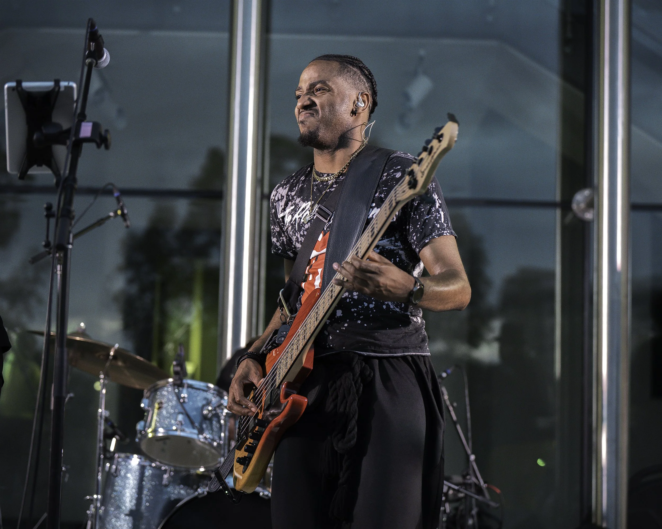 A musician playing an electric guitar during a performance, with a drum set in the background and microphone stand to the left.
