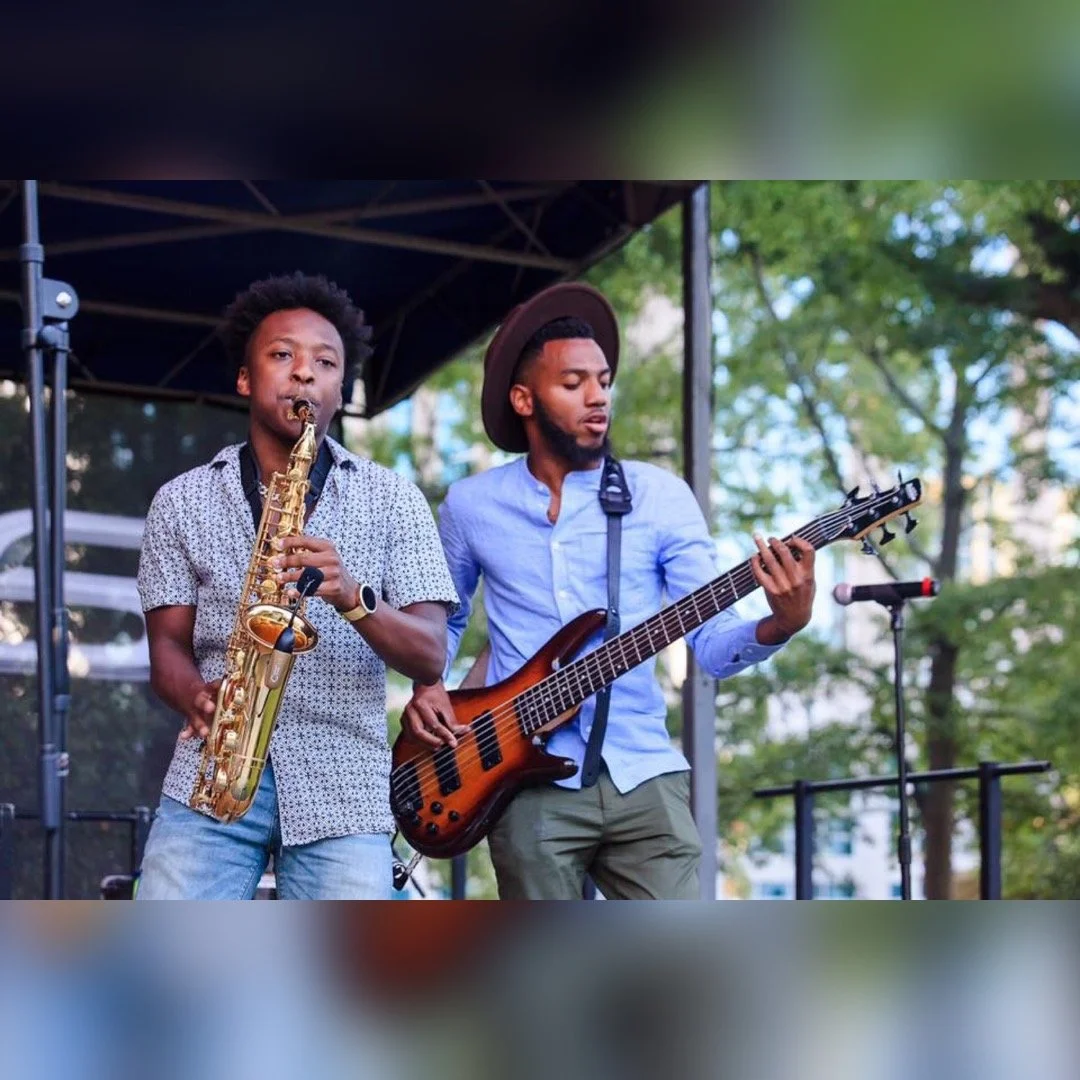 Two musicians playing guitars and saxophone on outdoor stage.
