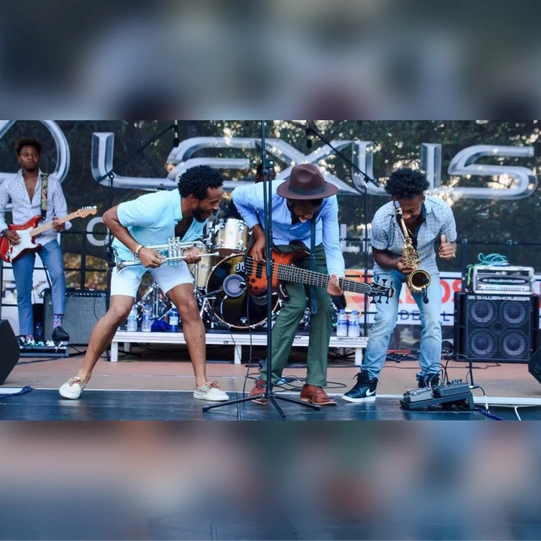 A group of five musicians performing on stage with various instruments, including an electric guitar, bass, saxophone, and drums, outdoors during daytime.