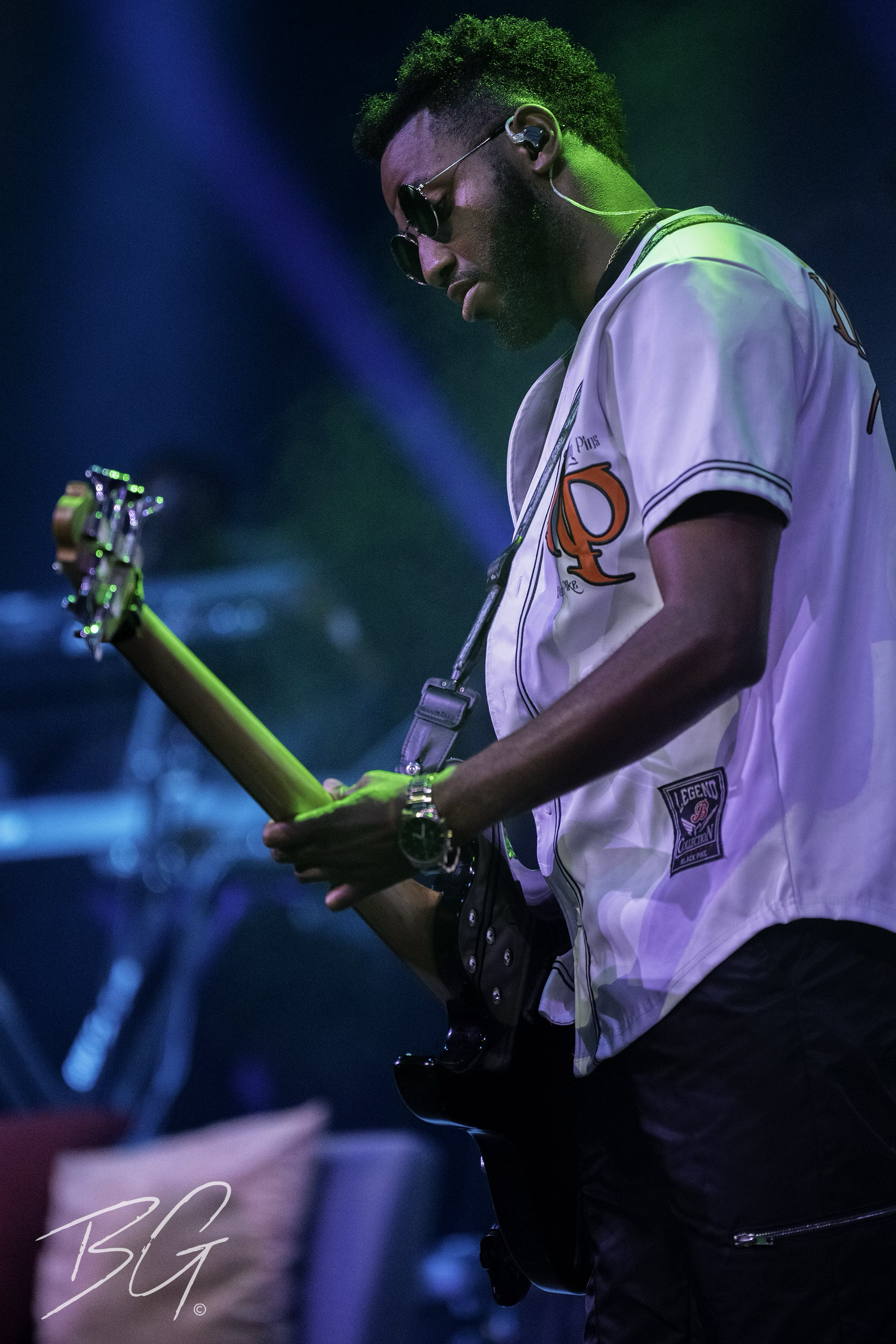 A man playing an electric guitar on stage, wearing sunglasses and a baseball-style jersey with a letter 'Q' on it, under colorful stage lighting.
