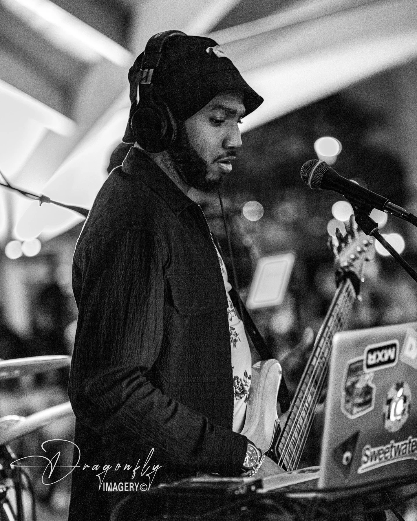 A young man with a beard, wearing a black beanie, headphones, and a dark jacket, is playing an electric guitar in a performance space. He is singing into a microphone and looking down at his guitar, with various stickers and labels on equipment in th