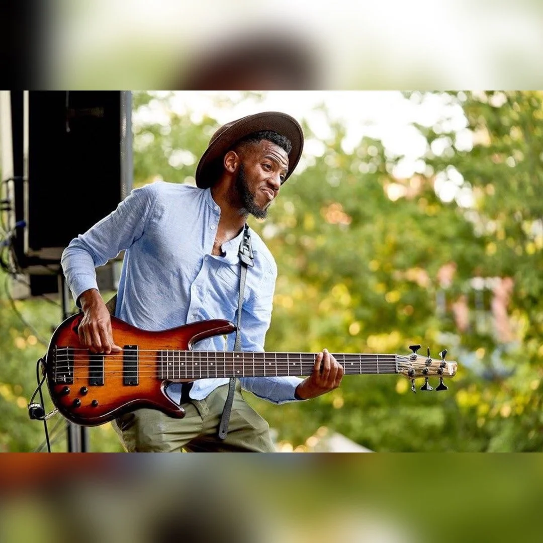 Man playing an electric bass guitar outdoors with trees in the background, wearing a hat and a light blue shirt.