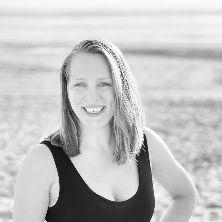 A smiling young woman with shoulder-length hair in a black sleeveless top standing on a beach.