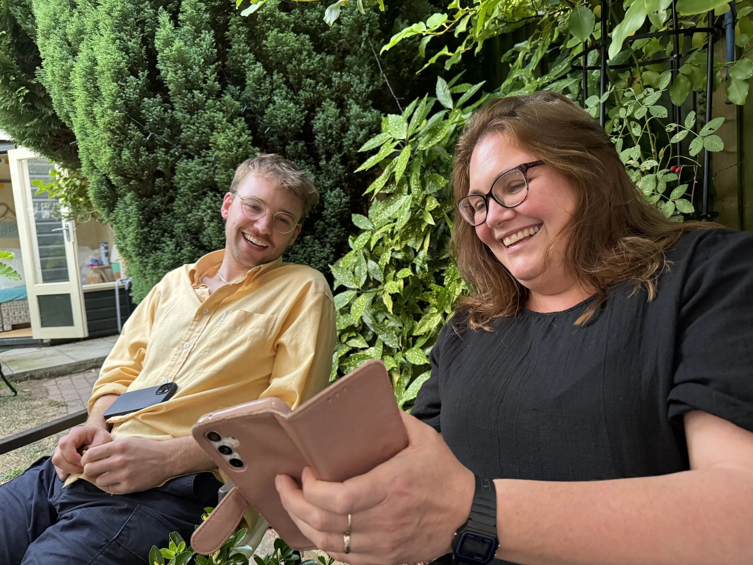 Two smiling people, a man and a woman, sitting outside in a garden with lush greenery. The woman is looking at her phone, and the man is looking at her, holding a smartphone.