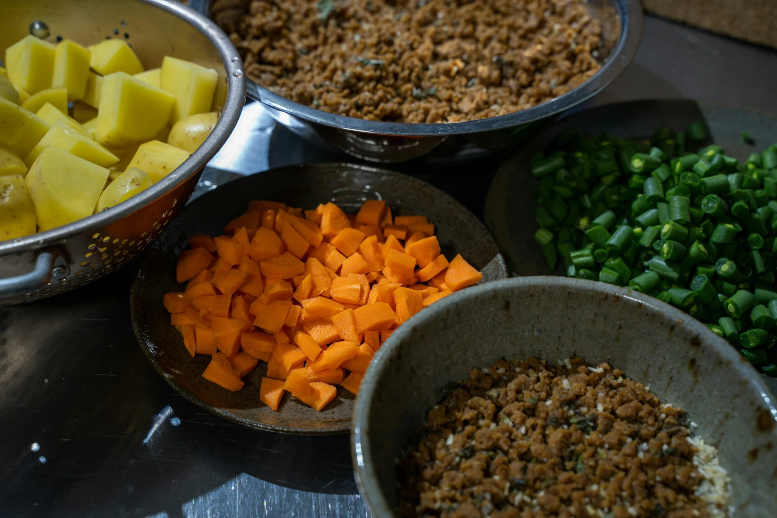 Assorted chopped vegetables including potatoes, carrots, green beans, and cooked ground meat in bowls on a kitchen counter.