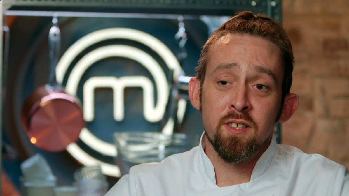 A male chef with brown hair and a beard speaking in front of a MasterChef logo in a kitchen.