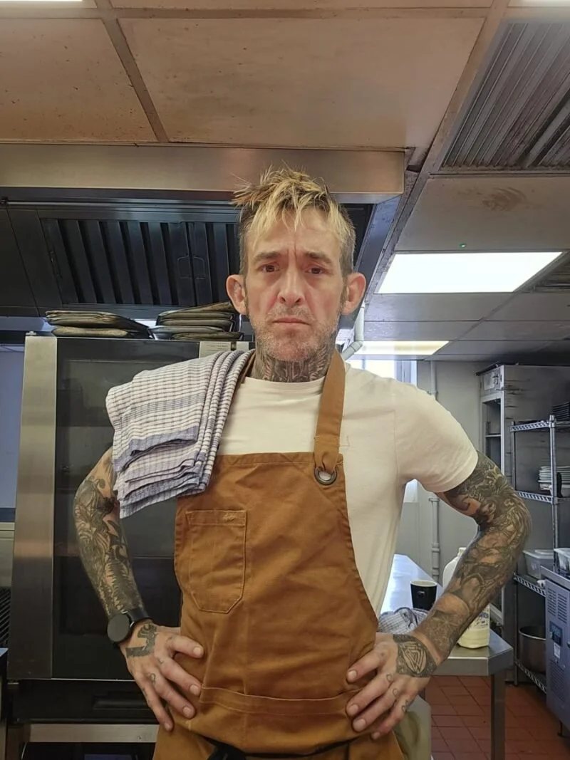A man with tattoos and messy blond hair stands with arms akimbo in a kitchen. He wears a white t-shirt and a brown apron, with a towel draped over his shoulder. The kitchen has industrial appliances and shelving in the background.