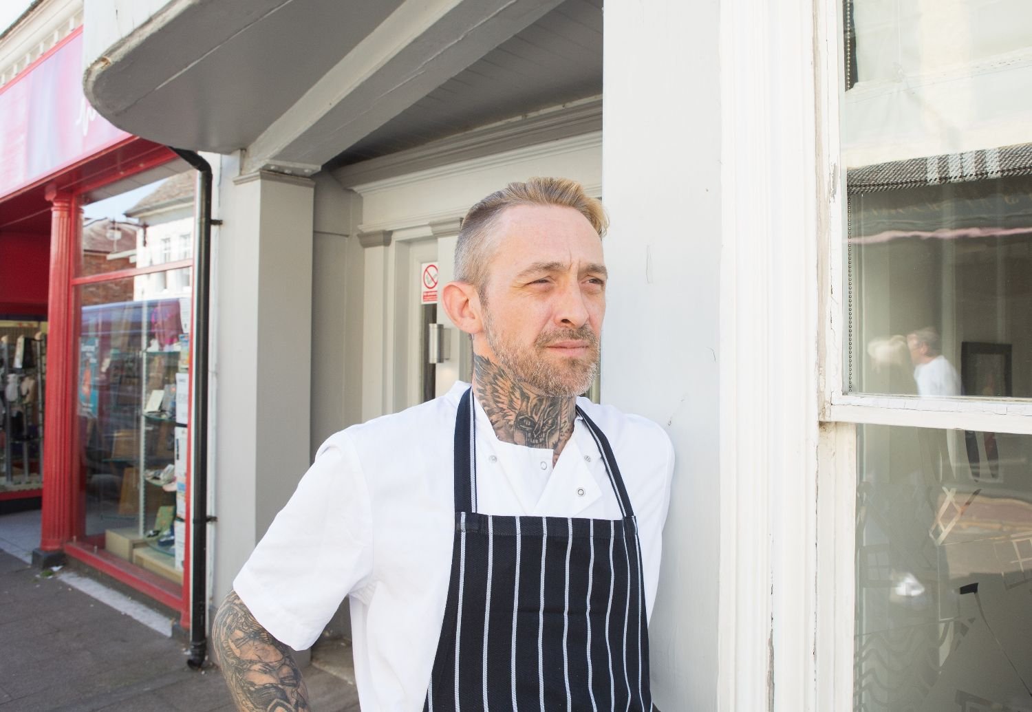 A man with tattoos on his neck and arms wearing a white chef's coat and a black and white striped apron, standing outside near a white building and a window with a reflection of a person.