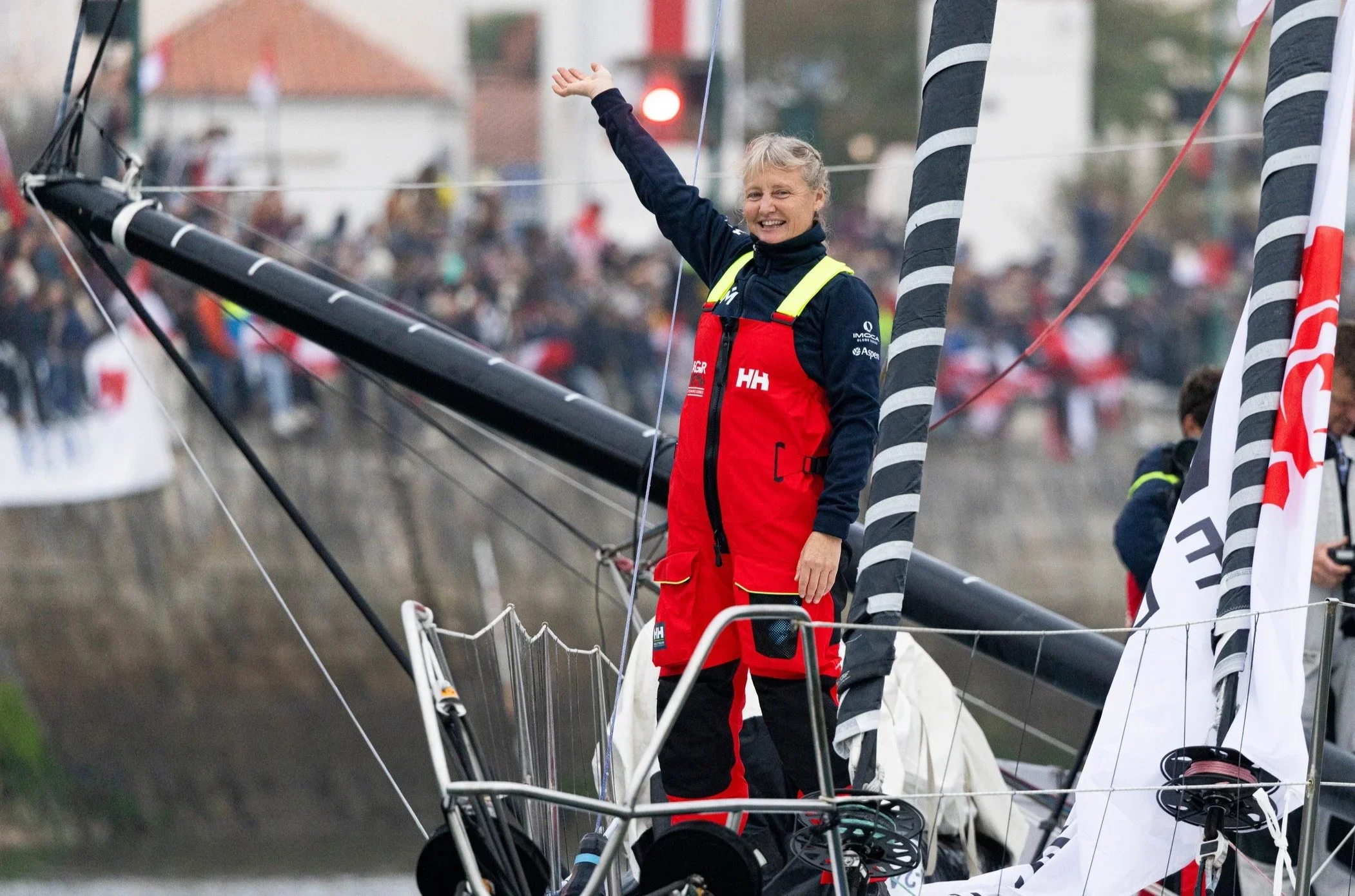 Pip Hare Ocean Racing sailor in red Helly Hansen sailing gear waving from the bow of a sailboat while motoring down the Les Sables-d’Olonne channel during the Vendée Globe.