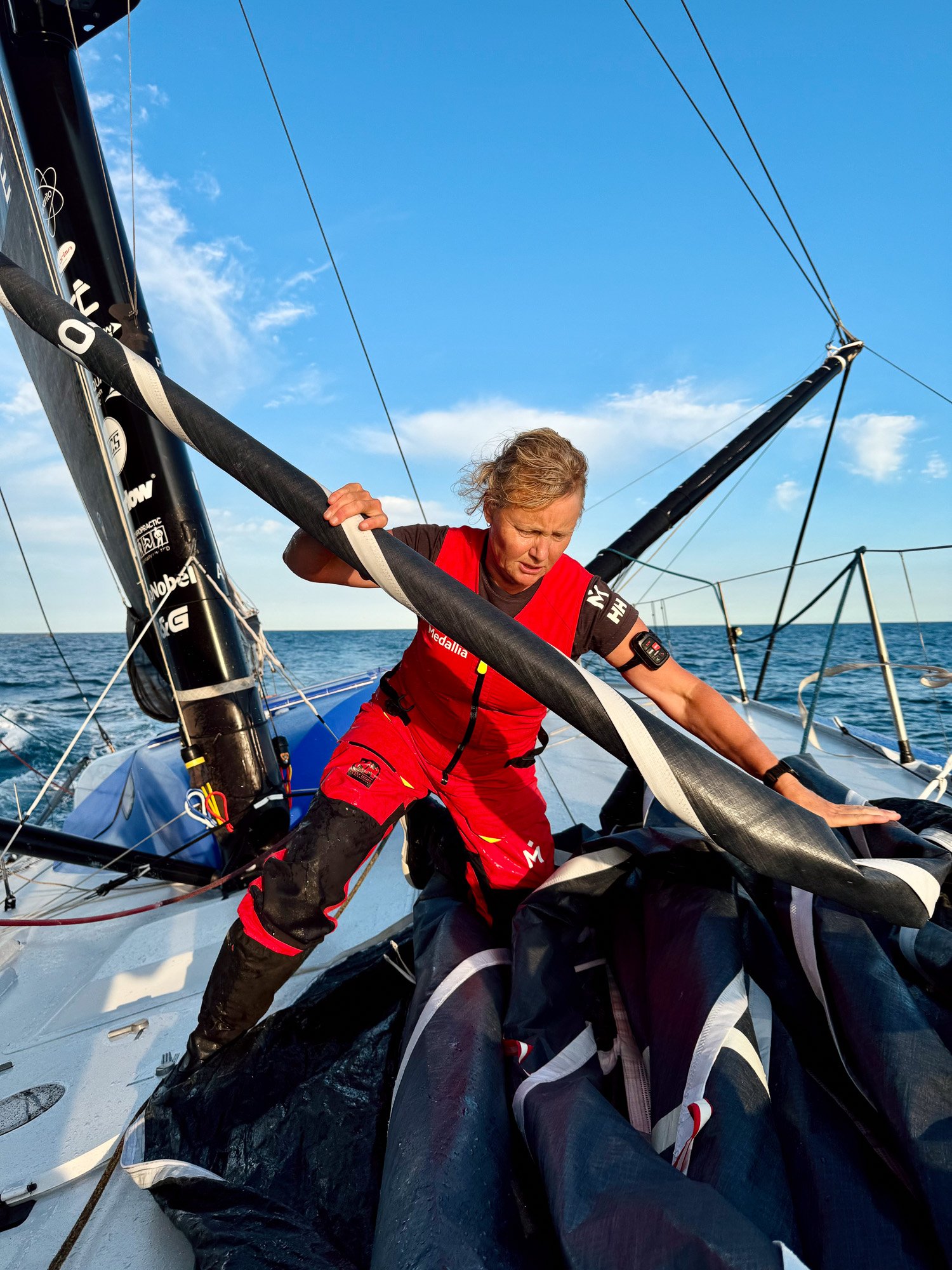 Pip Hare Ocean Racing - A woman in red sailing gear on a boat adjusting equipment, with the sea and a blue sky in the background.